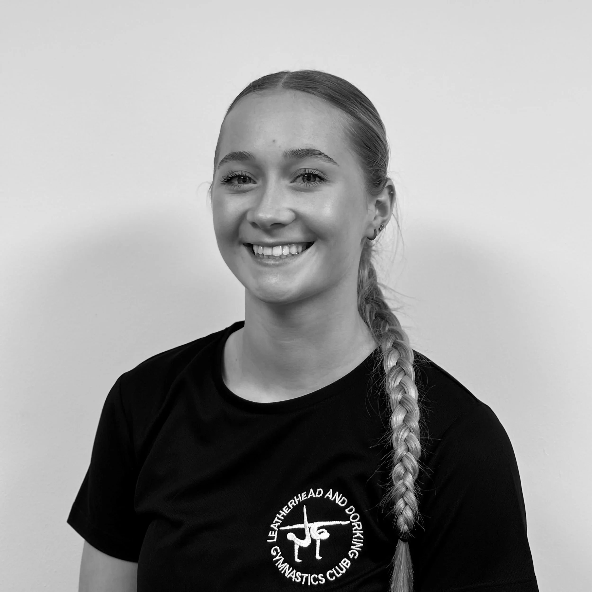 A young woman with braided hair smiling, wearing a dark T-shirt with white circular text around a logo, standing against a plain light-colored background.