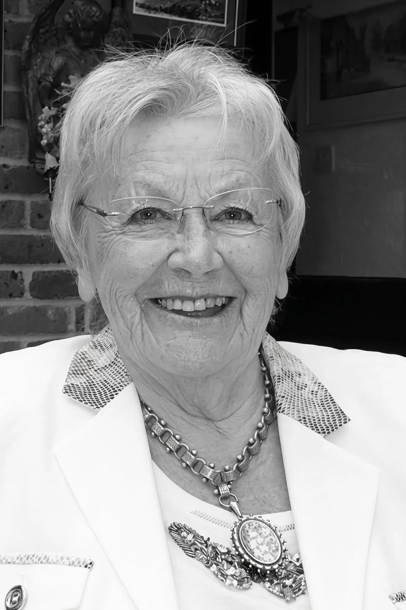 An elderly woman with short, light-colored hair smiling, wearing glasses, a white blazer, and a decorative necklace, seated indoors with brick wall and photographs in the background.