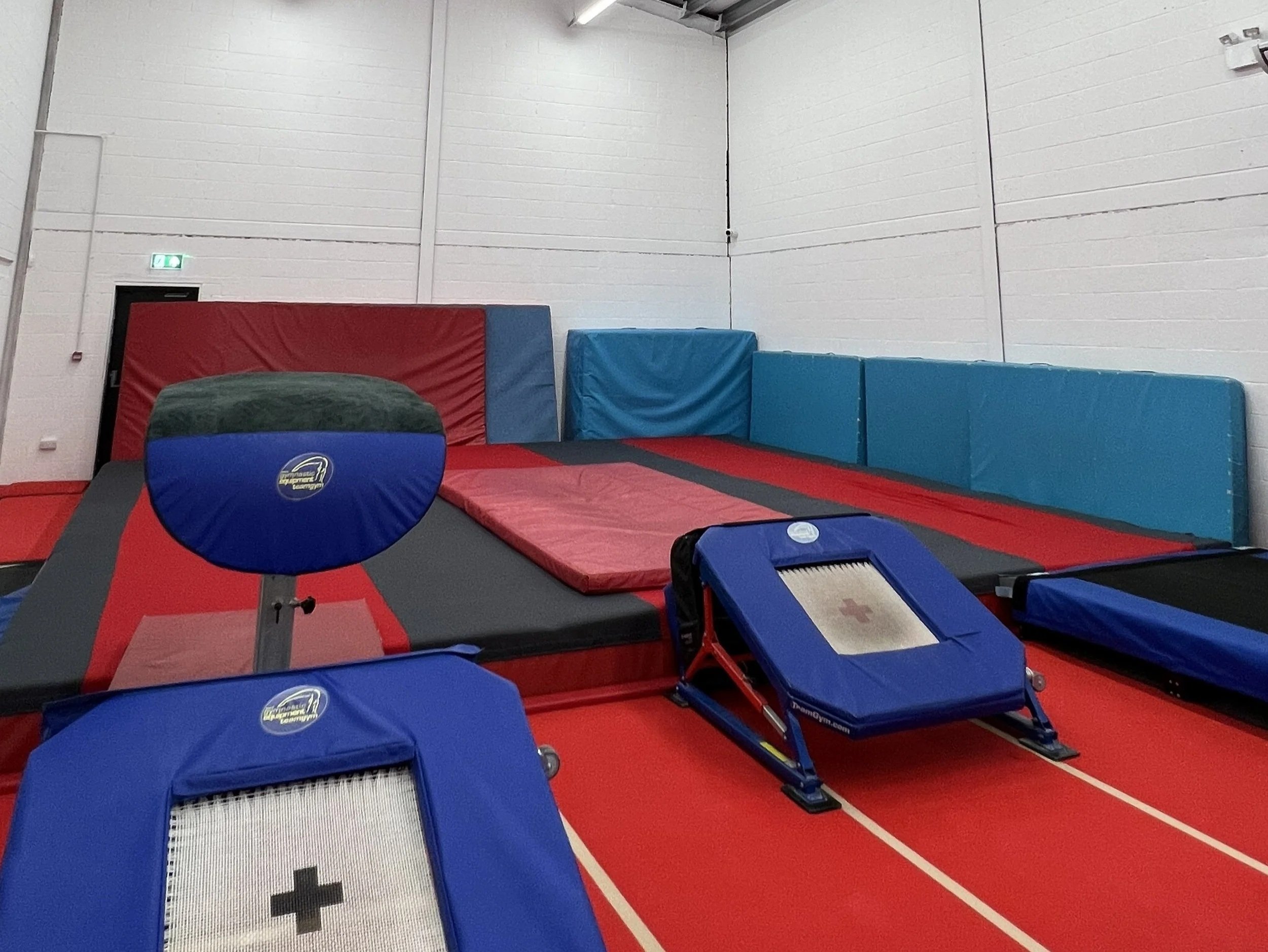 Indoor gymnastic training area with red and blue padded mats, a trampoline, and foam blocks against white brick walls.