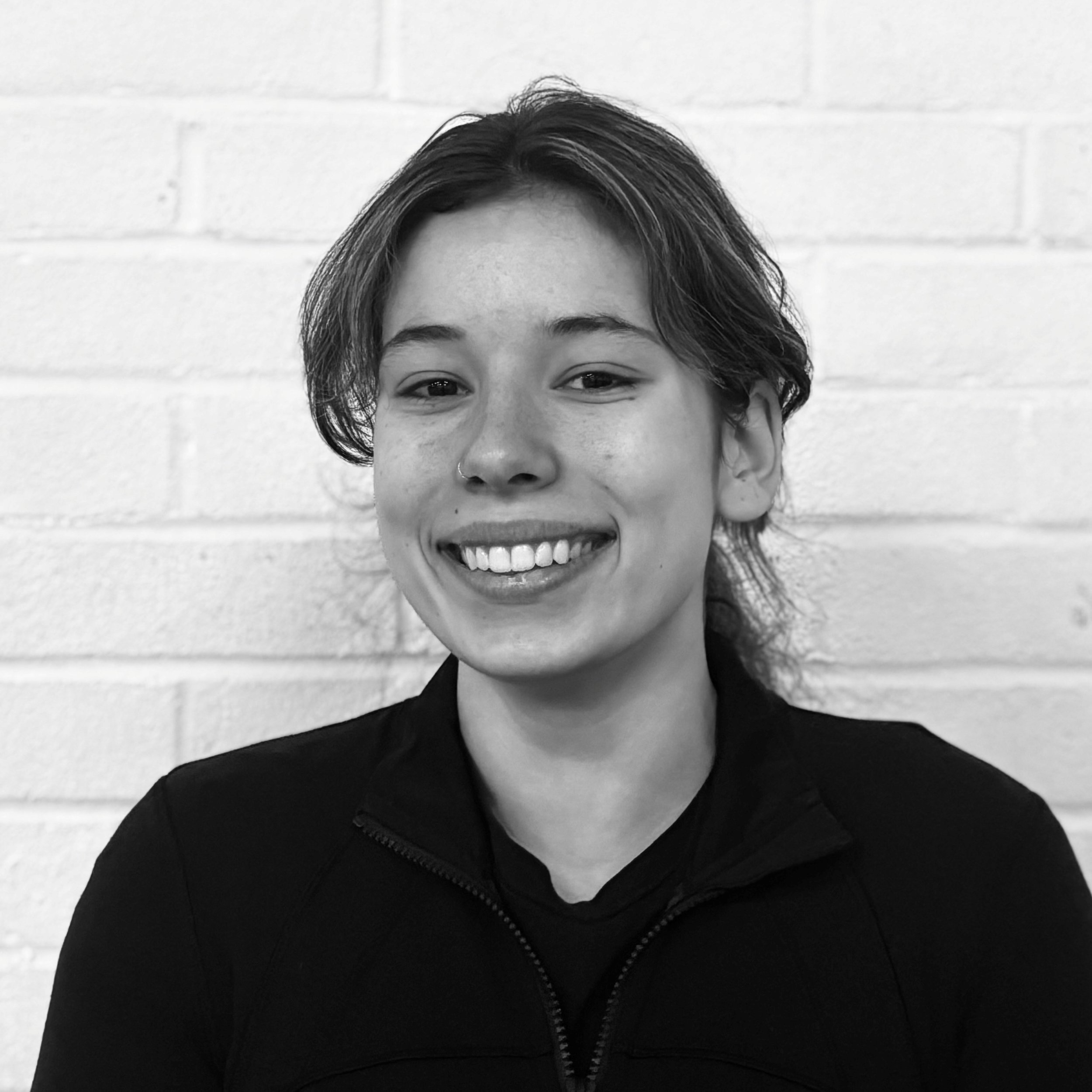 Close-up black and white photo of a smiling person with short hair, wearing a black zip-up jacket, standing against a white brick wall.