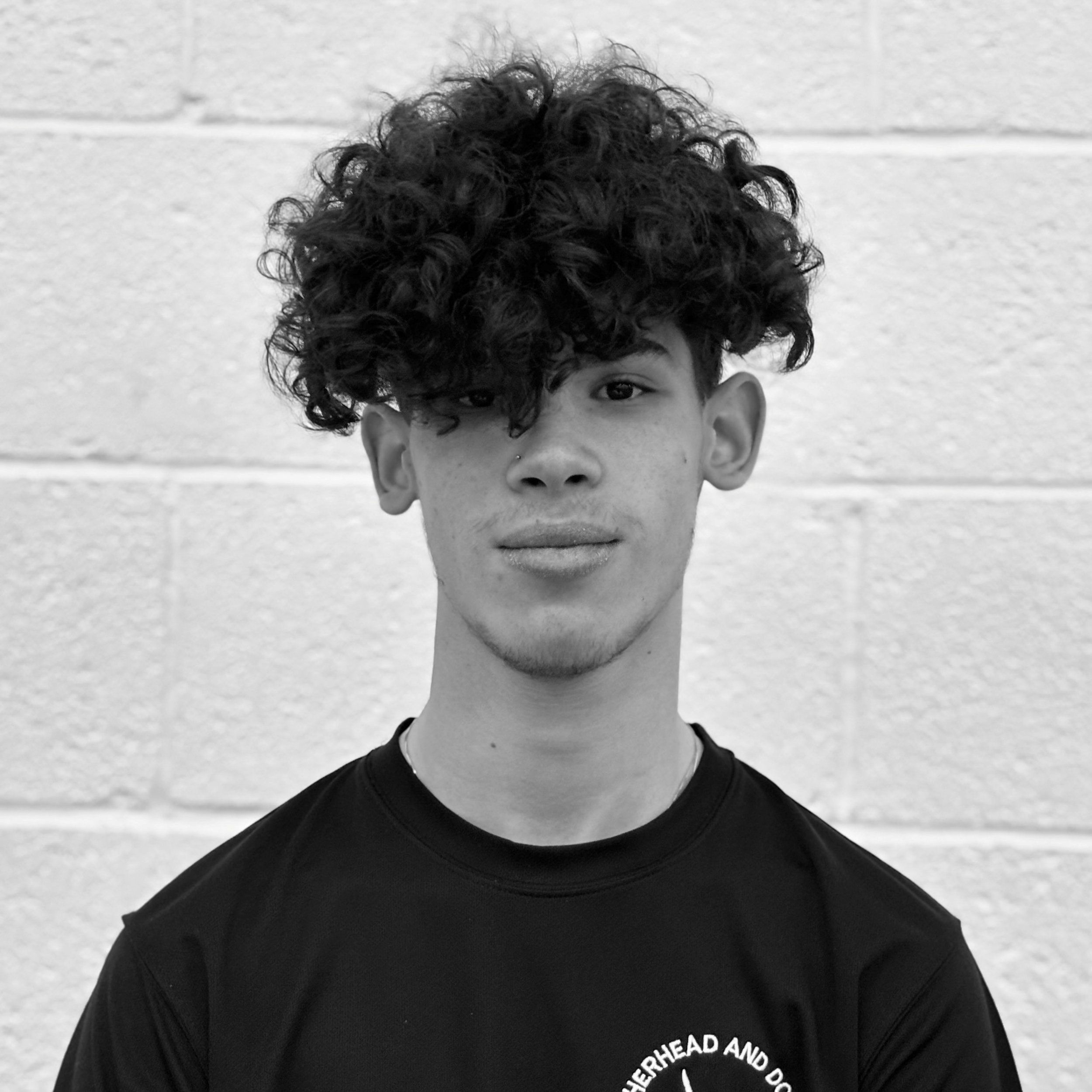 Close-up of a young man's face with curly hair, wearing a black t-shirt, standing outdoors against a background of a concrete wall.
