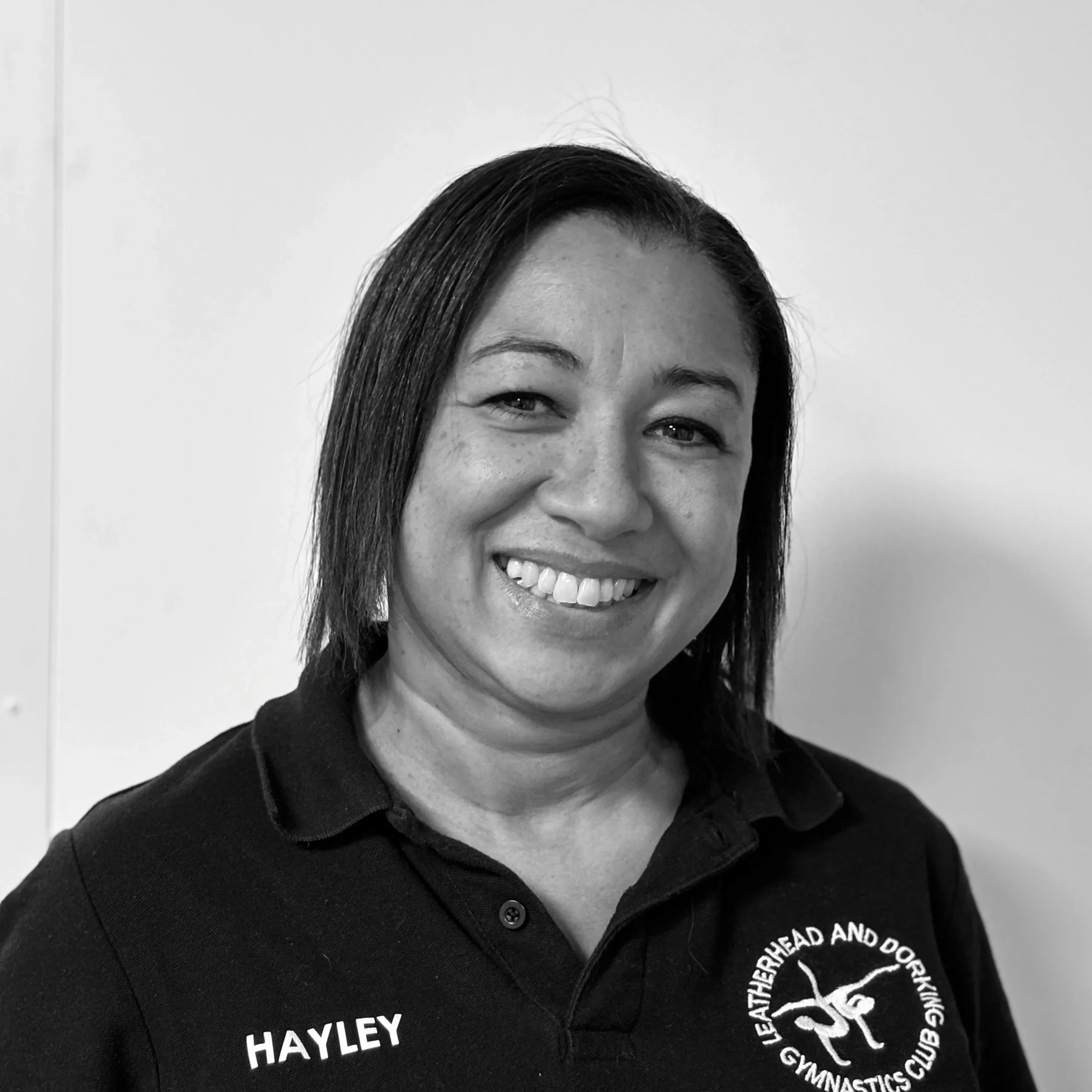 Close-up of an Asian woman with straight hair smiling, wearing a black polo shirt with text and a circular logo for a gymnastics club.
