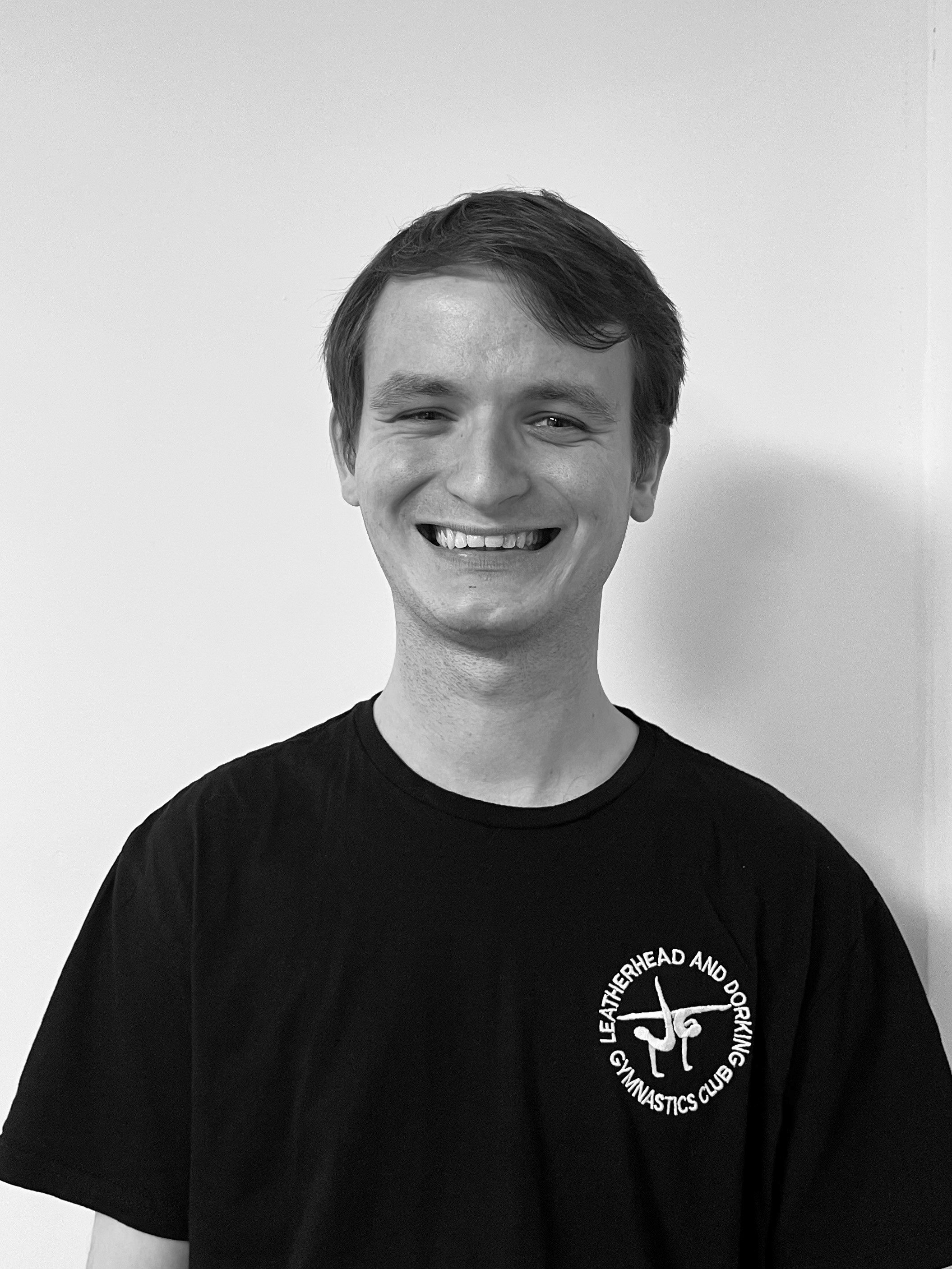 Black and white photo of a young man smiling, wearing a black t-shirt with white text and a logo related to a gymnastics club, standing against a plain light-colored background.