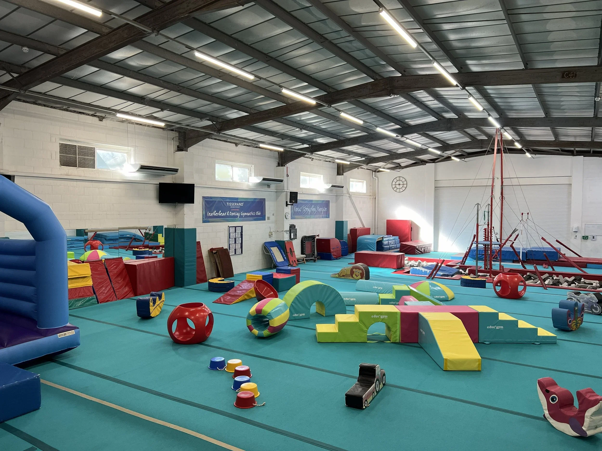 Indoor gymnastics training area with colorful foam blocks, climbing equipment, and padded mats on a green gym floor. The space has white brick walls, high ceiling with exposed beams, and windows letting in natural light.