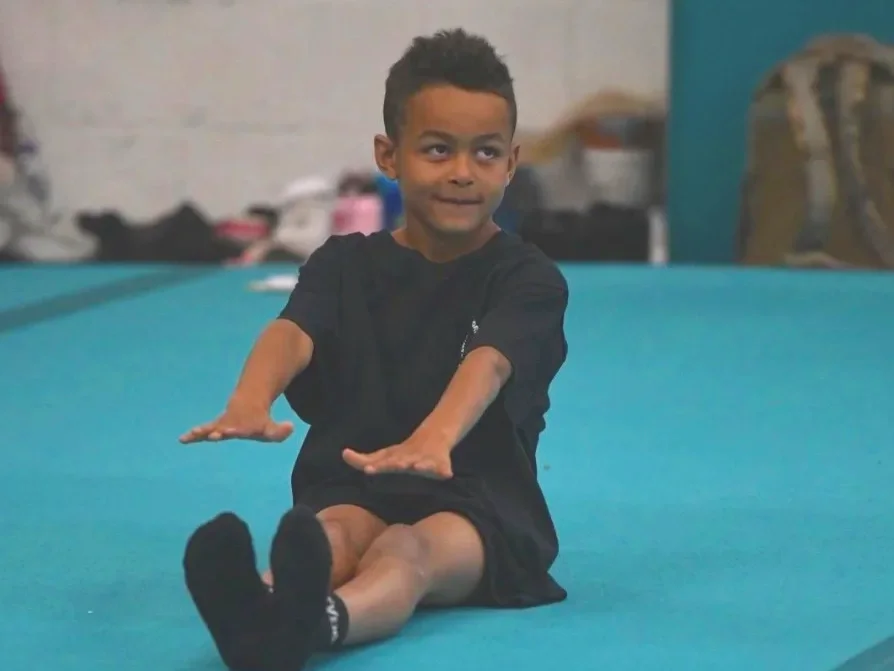 A young boy wearing glasses and a purple T-shirt with science and gymnastics text extending his arms outwards, possibly practicing balance or gymnastic poses at an indoor gym or sports event.