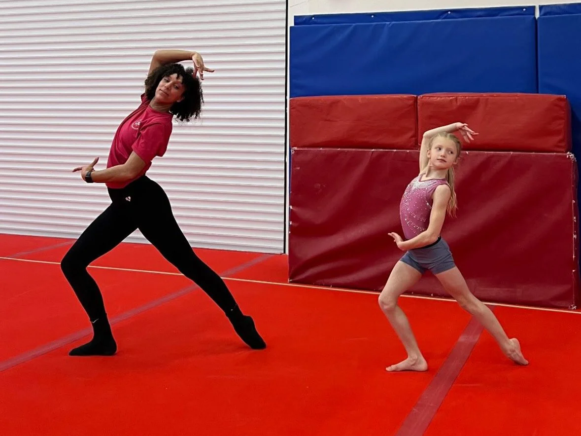 A woman and a young girl practicing dance or gymnastics in a gym with red mats, red and blue padded walls, and a white rolling door.