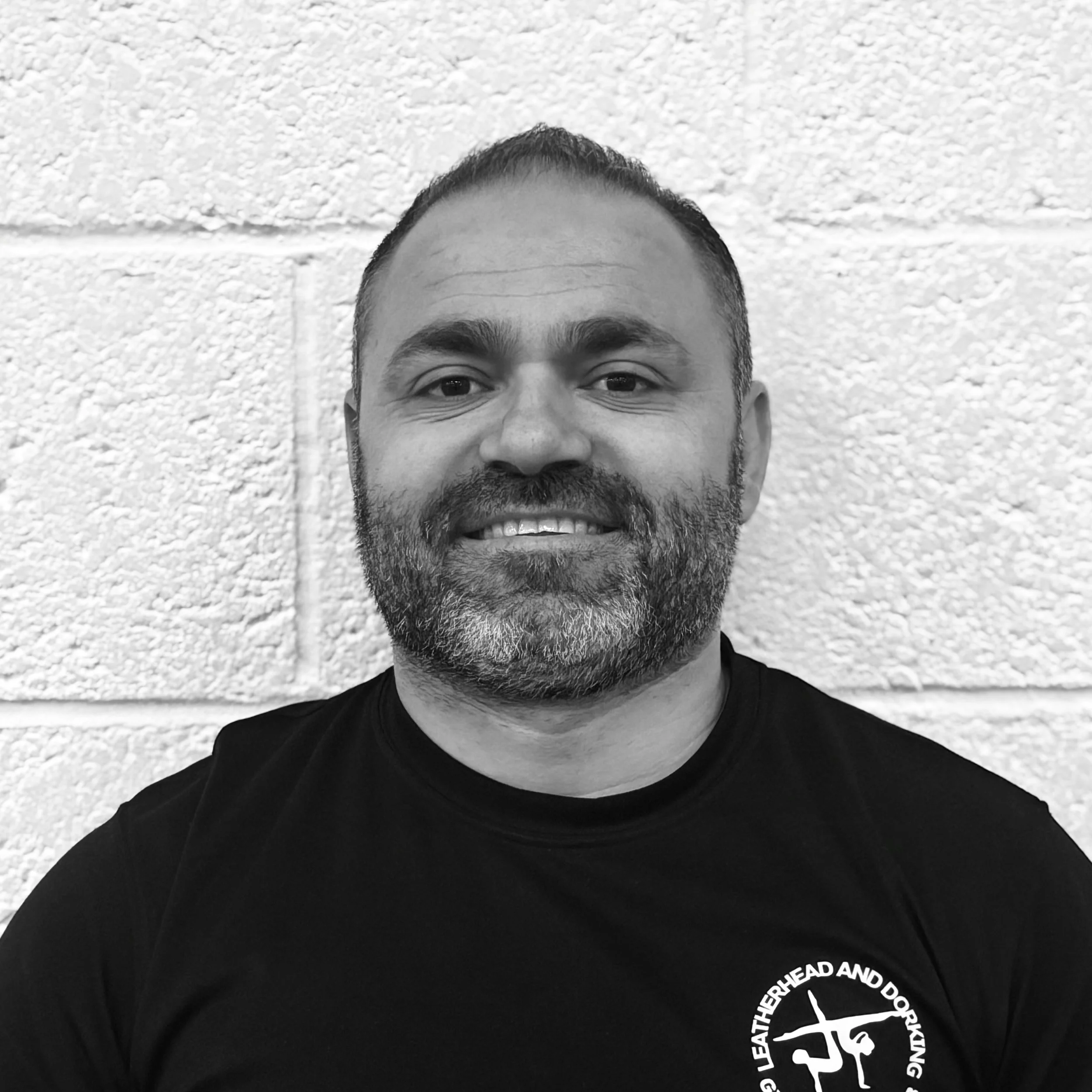 Close-up of a man with short dark hair and a beard, smiling against a textured white wall wearing a black t-shirt with white text and a logo.