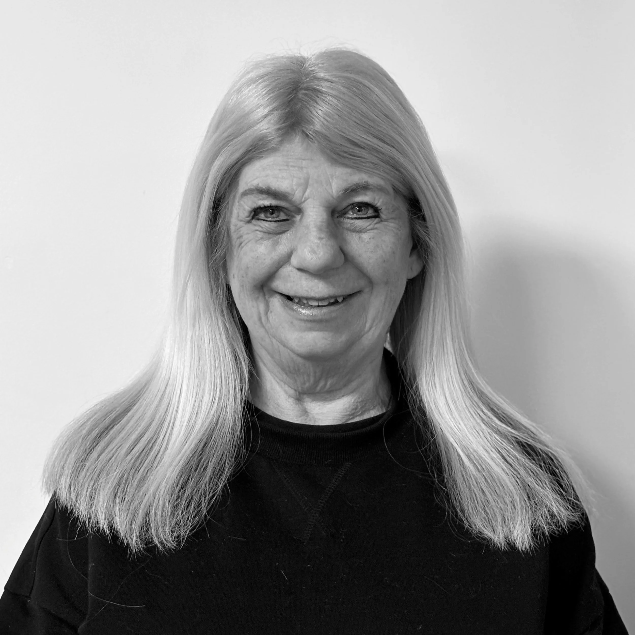 Close-up of a smiling woman with long gray hair and blue eyes, wearing a black top and a jacket with a fur collar, against a plain light background.