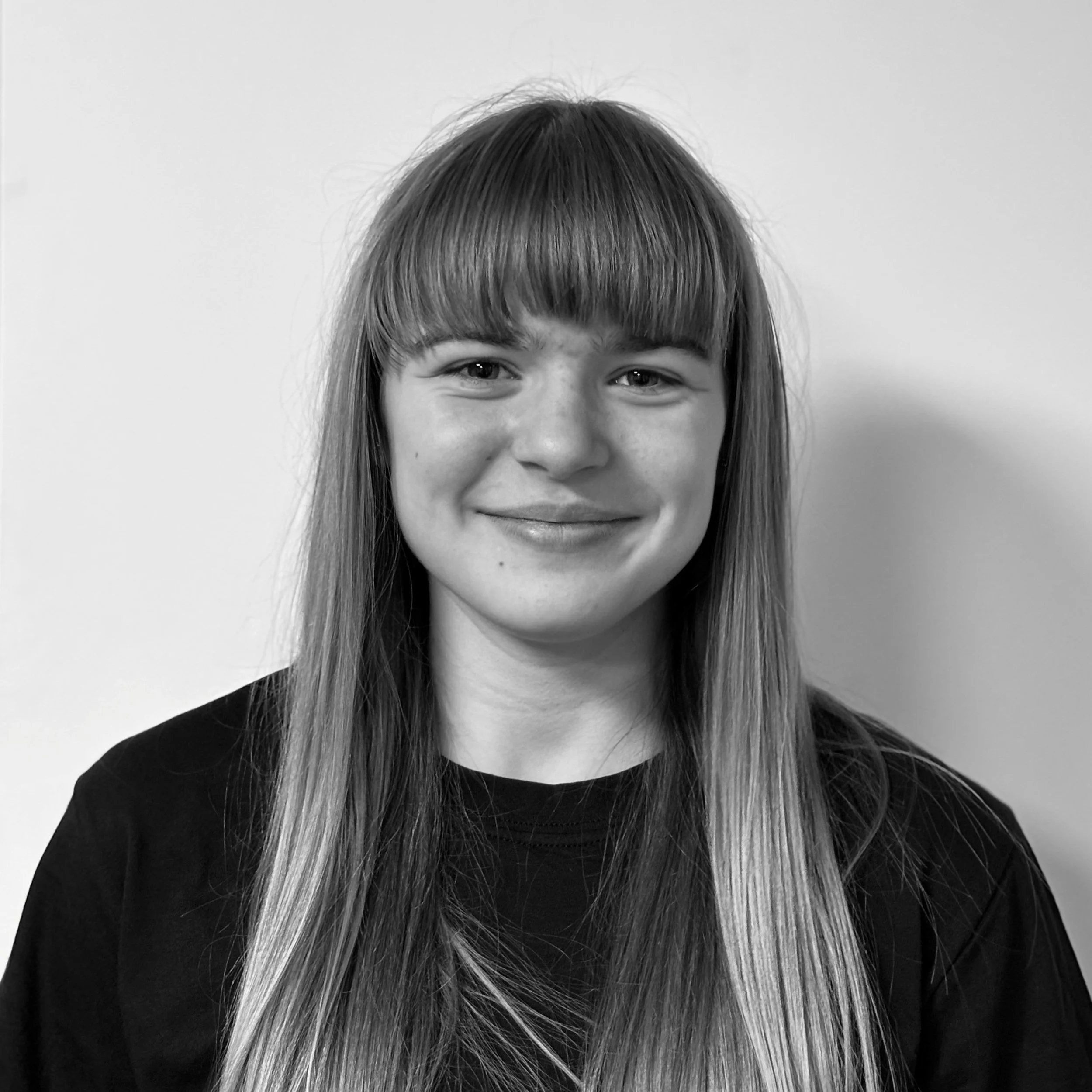 Black and white photo of a young girl with long hair and bangs, smiling, against a plain white background.