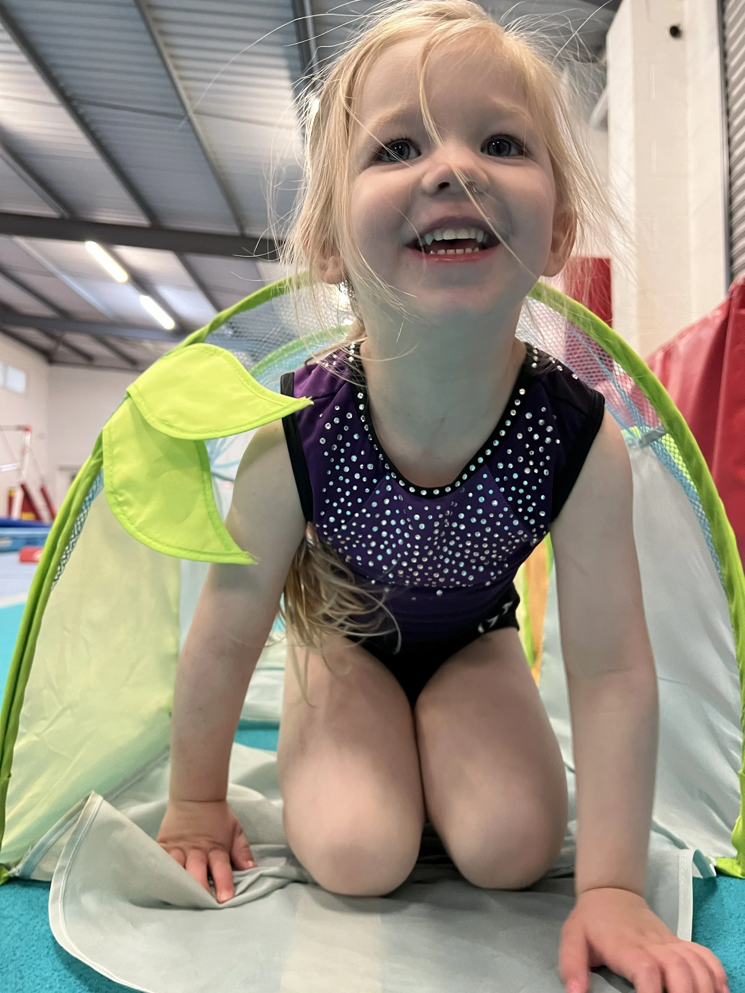 A smiling young girl with red hair and blue eyes crawling on the floor inside a gym or indoor play area, wearing a purple and black outfit, with a green and yellow netted cape around her shoulders.