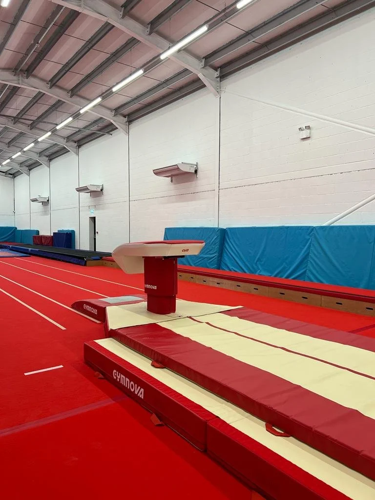 Indoor gymnastics training area with a red springboard and vaulting apparatus on a red mat, blue padded walls, and high ceiling with exposed beams and lighting.