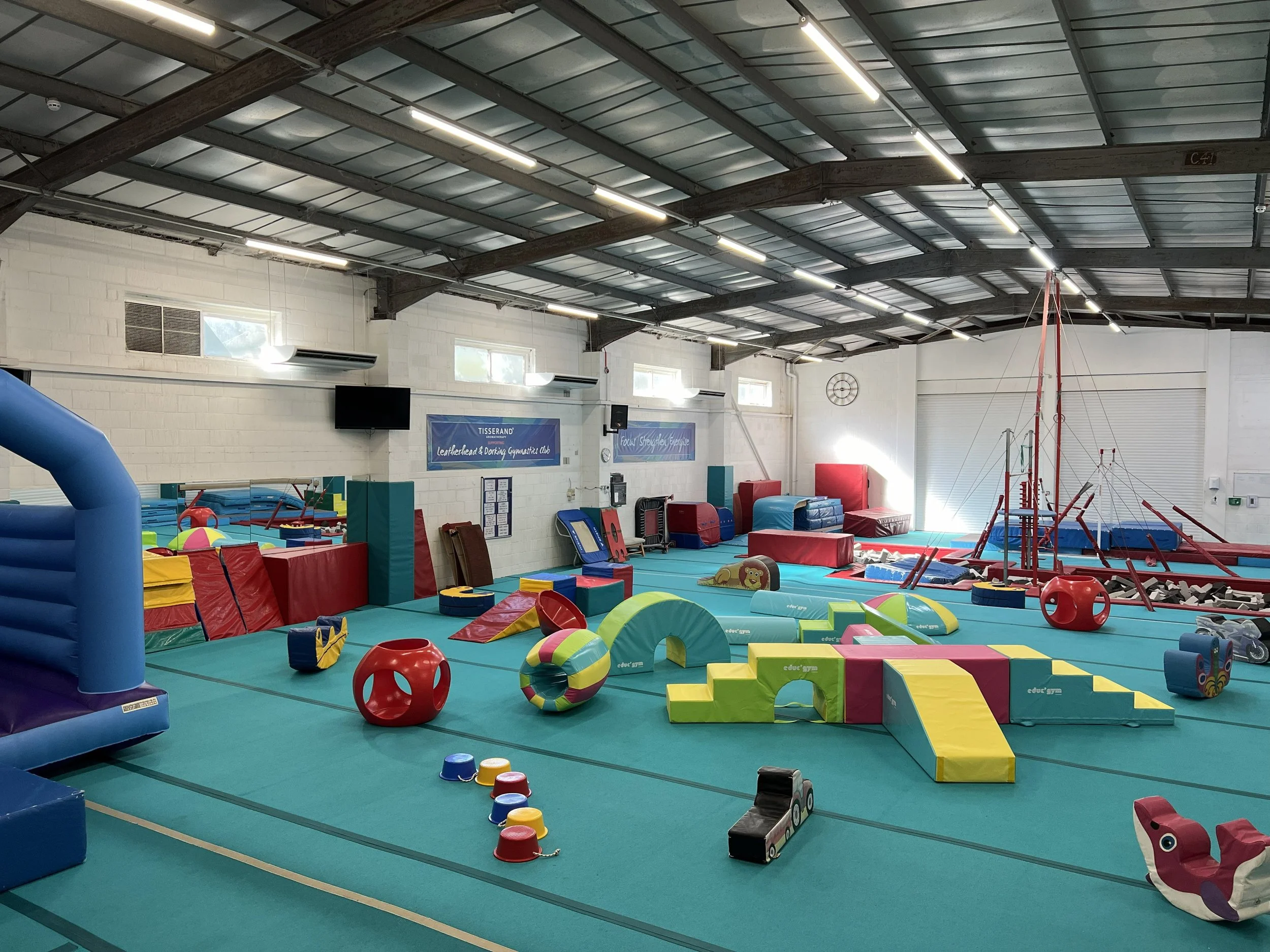 Interior of a gymnastics gym with various colorful mats, foam blocks, and climbing equipment arranged on a teal gym floor. The walls are white with windows and a flat-screen TV, and the ceiling has exposed beams and lights.