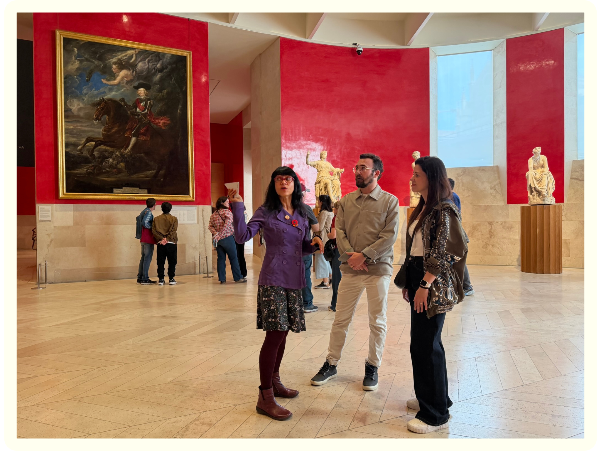 People observing artwork and sculptures in a museum with a large painting of a horse and rider on a red wall.