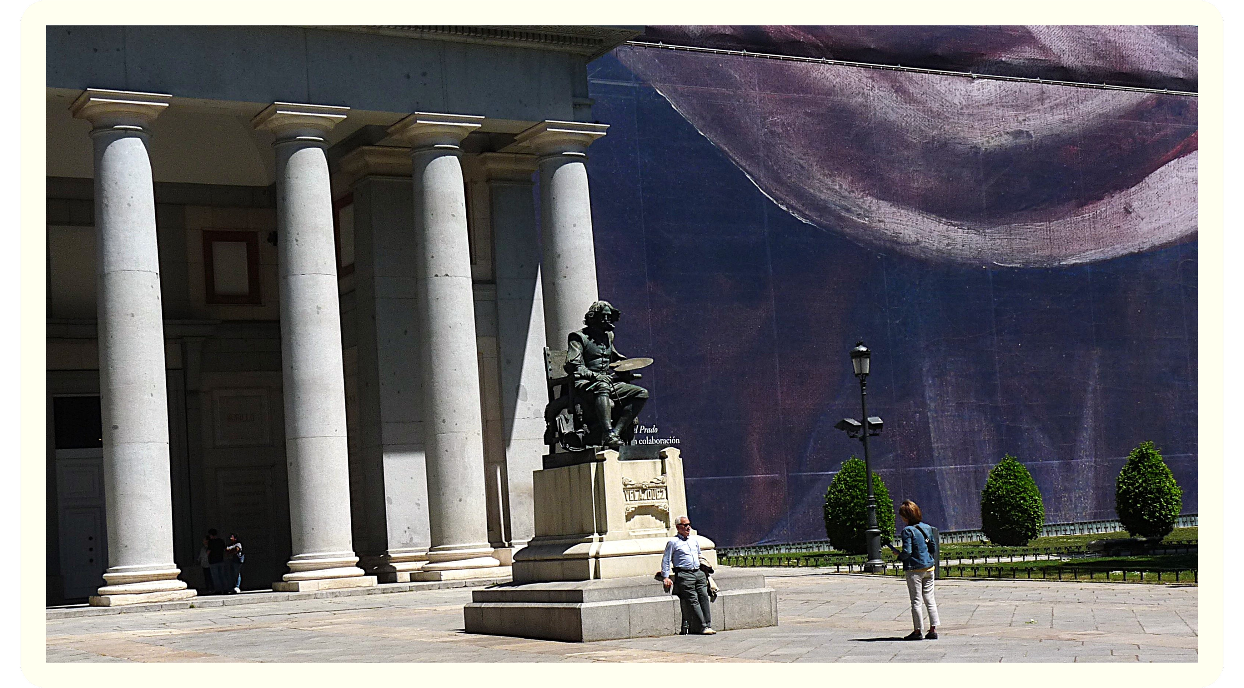 A large stone building with tall columns and a statue of a seated man holding a book in front of it. Several pedestrians are walking nearby. In the background, a mural or large artwork is visible on a wall.