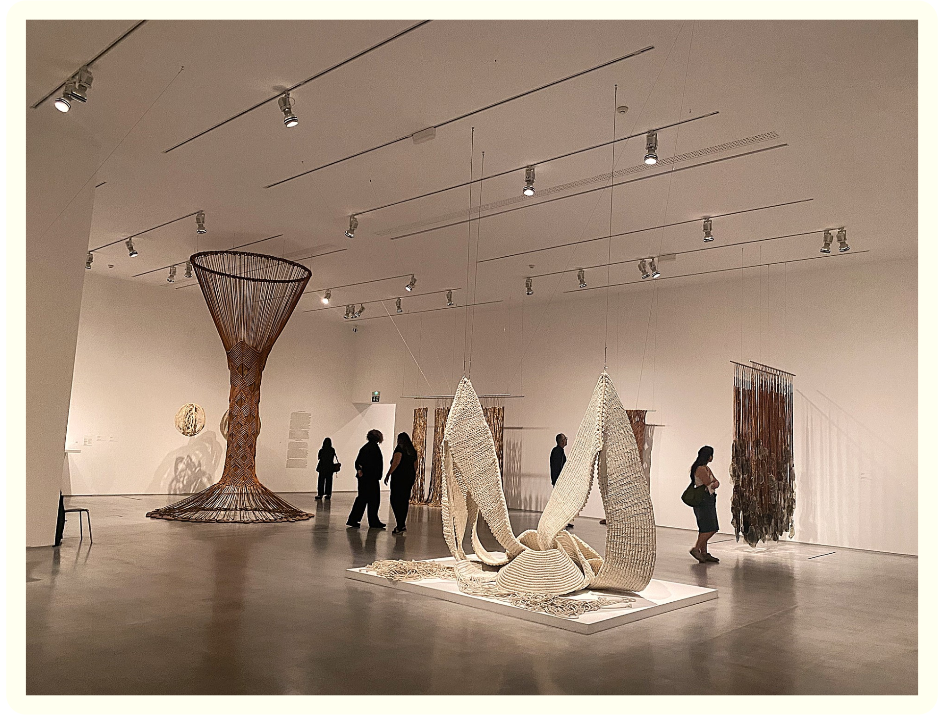 An art exhibit featuring large, intricate woven sculptures and textile installations, with visitors observing the artwork in a spacious gallery.