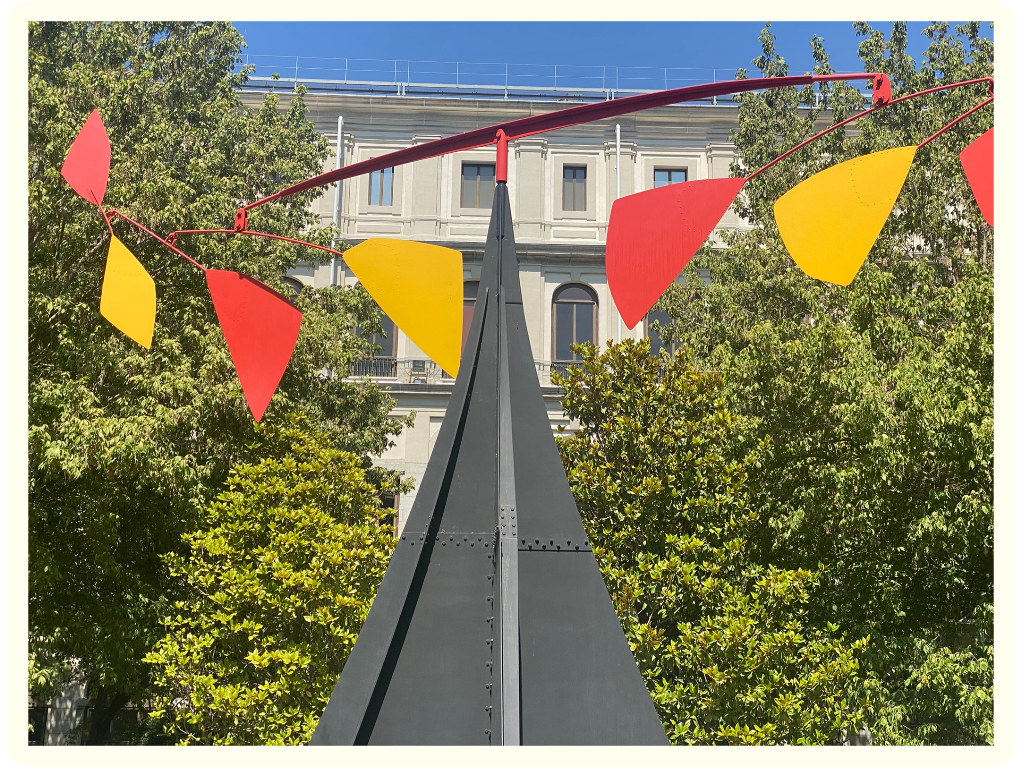 Photo of a decorative sculpture with red and yellow triangular shapes, set against green trees and a building with classical architecture, under a clear blue sky.