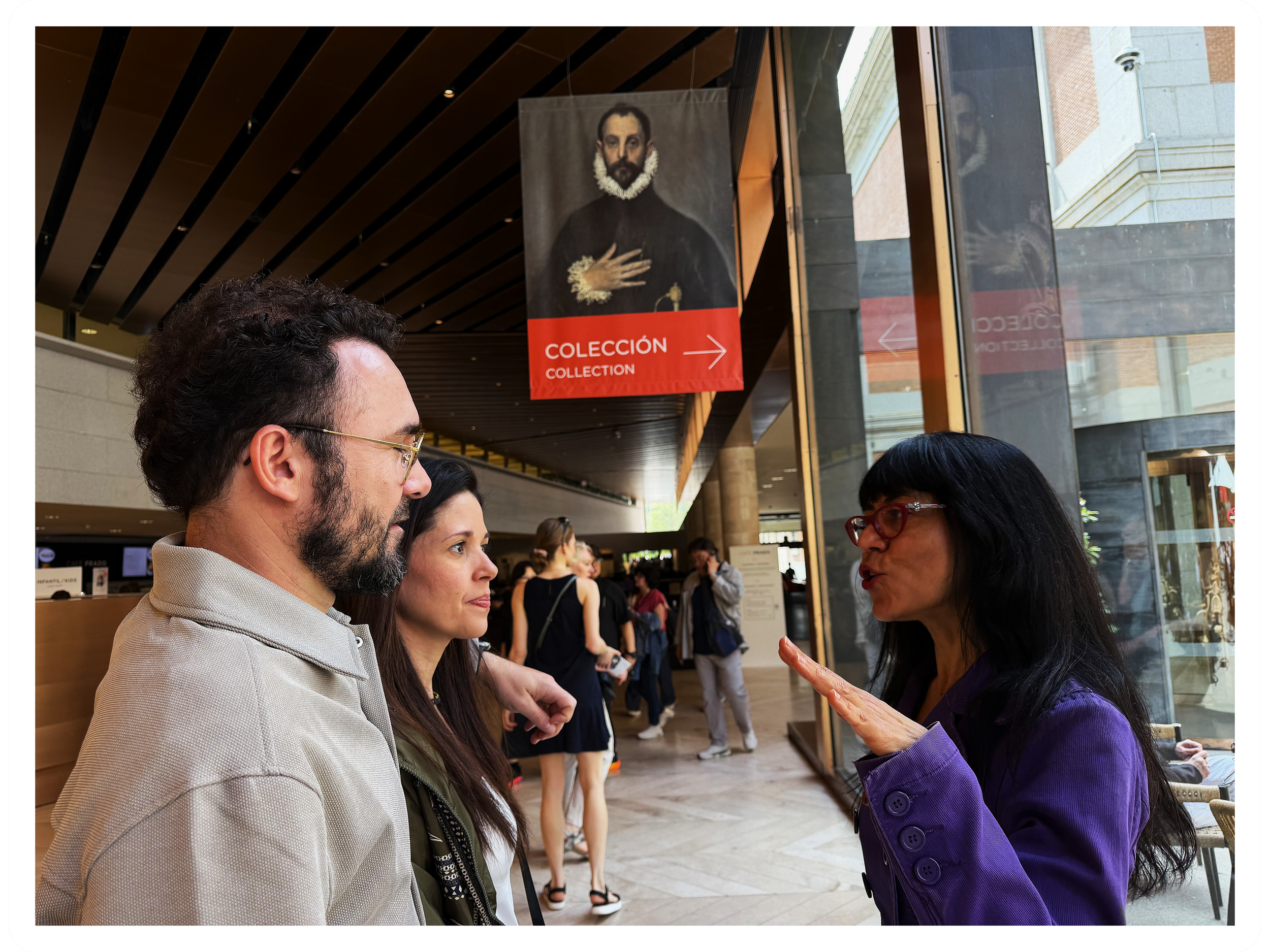 People lined up in a museum or gallery, engaging in conversation near a sign indicating a collection, with a woman in purple jacket speaking to a man with glasses.