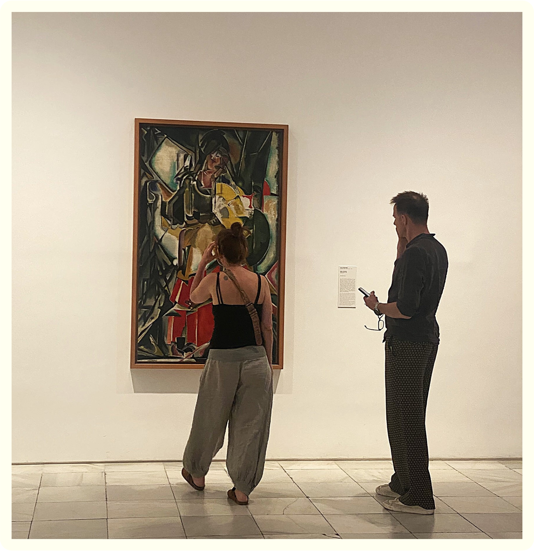 Two people are standing and looking at a colorful modern art painting in an art gallery or museum. The woman on the left has her hair tied up and is looking at the painting, while the man on the right is looking at his phone.
