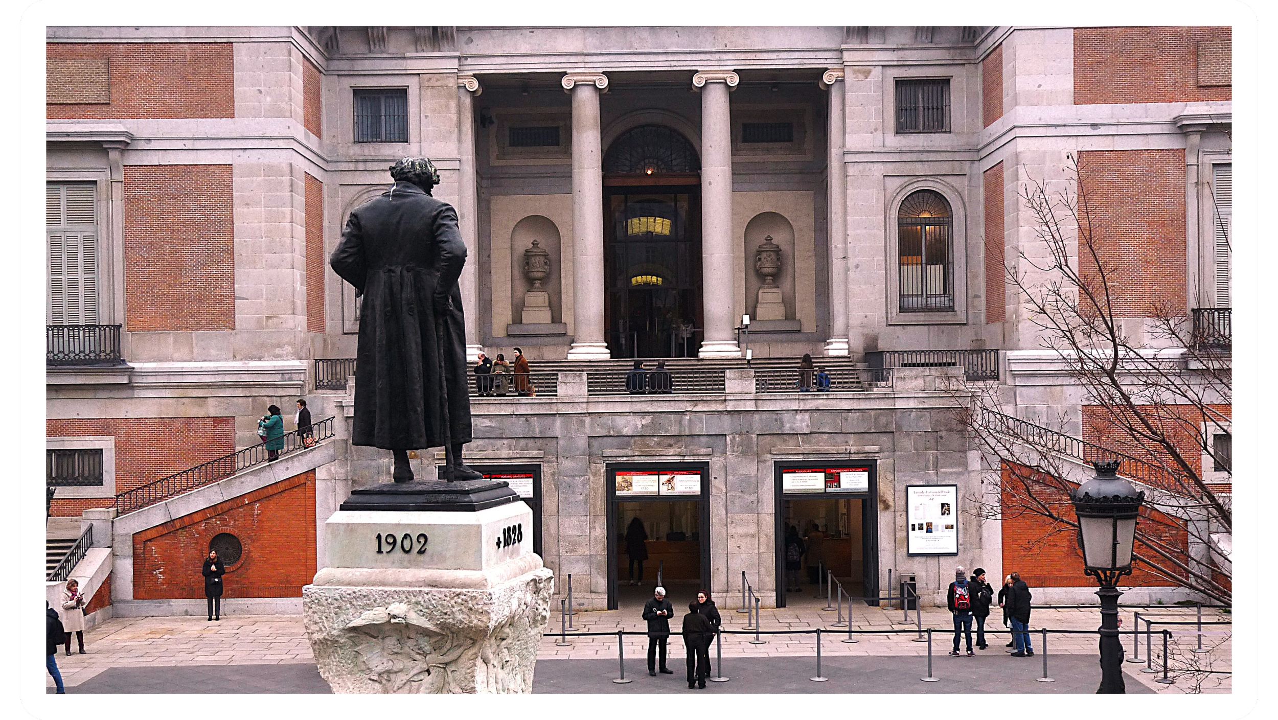 A group of people gathered outside the American Folk Art Museum in New York City. The building features large columns, brick and stone facade, and large windows. There is a statue of a man with a coat and curly hair in the foreground and a few leafle