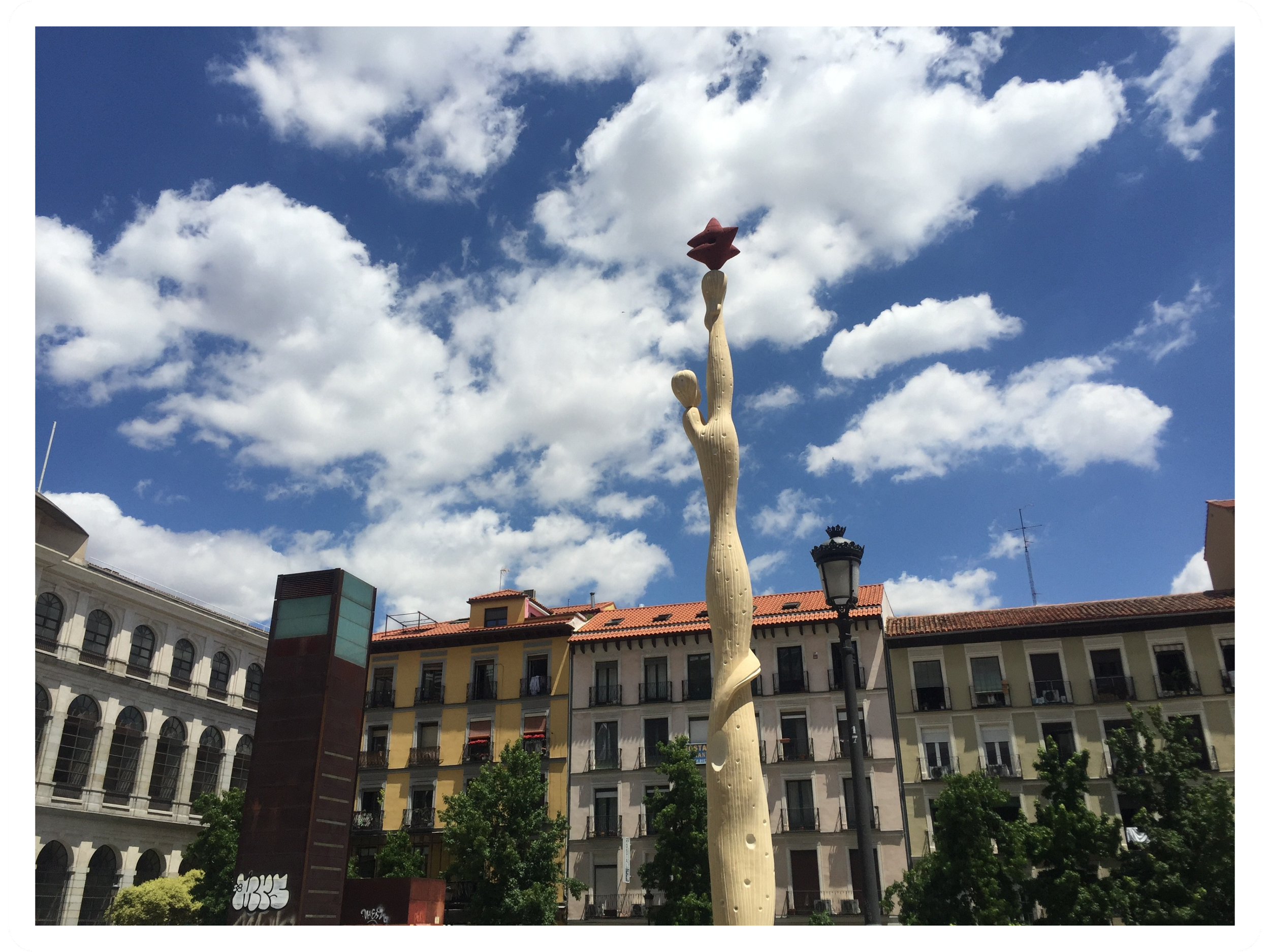 A tall sculpture resembling a cactus with a pink star-shaped top, set against a partly cloudy blue sky, in an urban city square surrounded by buildings.