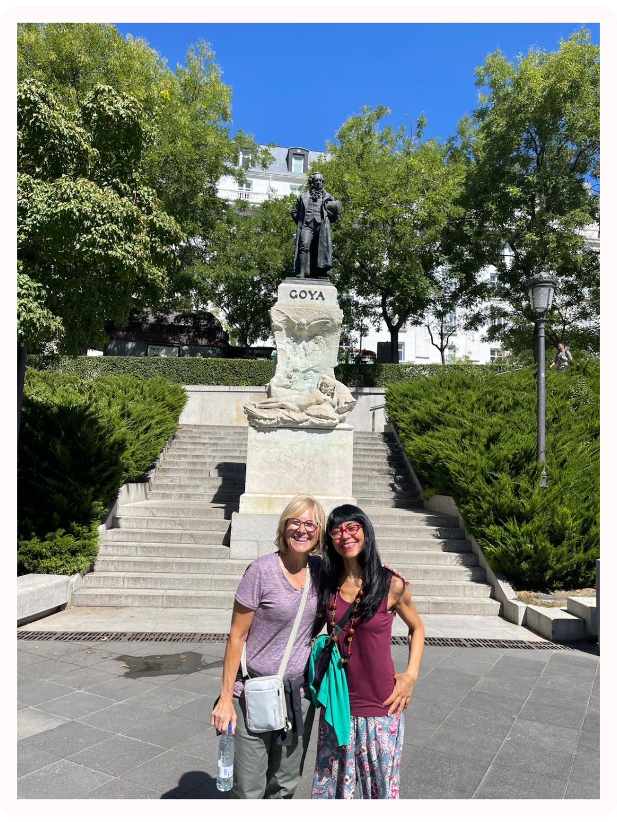 Two women standing in front of a statue of a man on a pedestal with sculptures of lions and a reclining figure below, at the Goya monument in a park surrounded by trees and greenery on a sunny day.