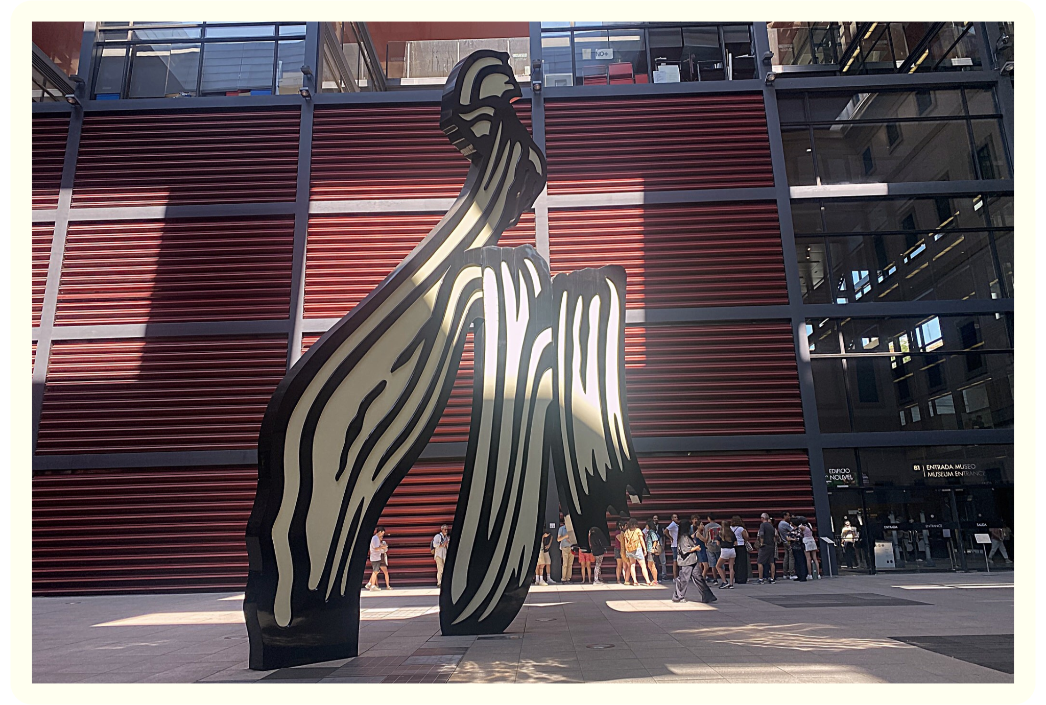A large black and white striped horse sculpture in front of a modern glass building with red horizontal panels, with people lining up outside the museum entrance.