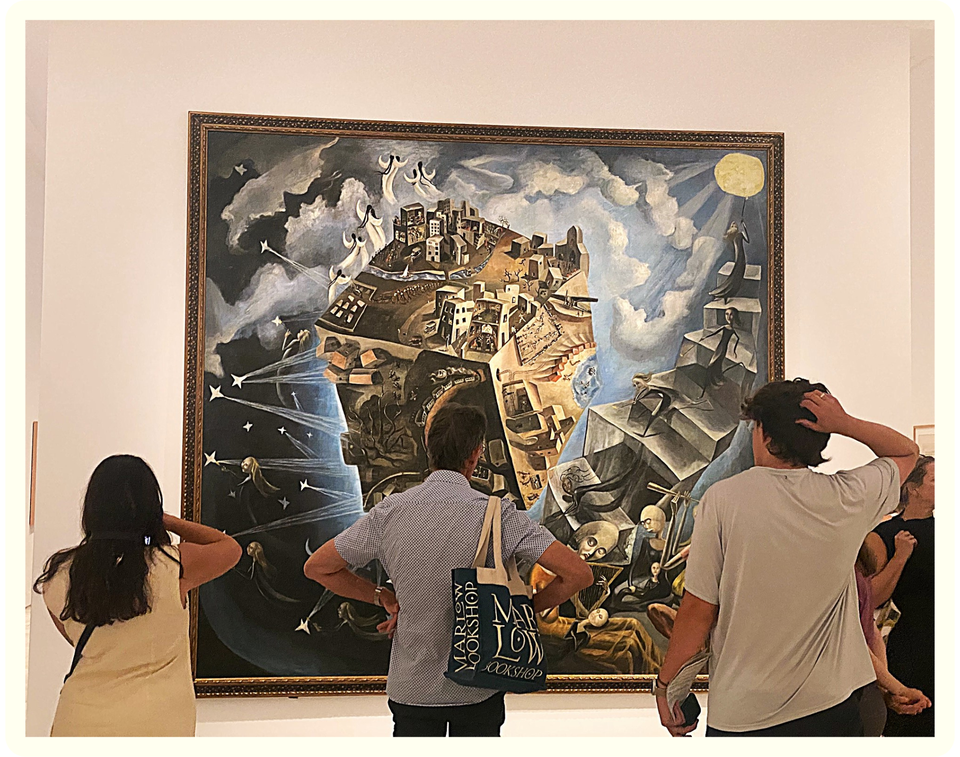 People viewing a large surrealist painting in an art gallery. The painting depicts a floating landscape with houses, figures, and celestial elements like moon and stars.