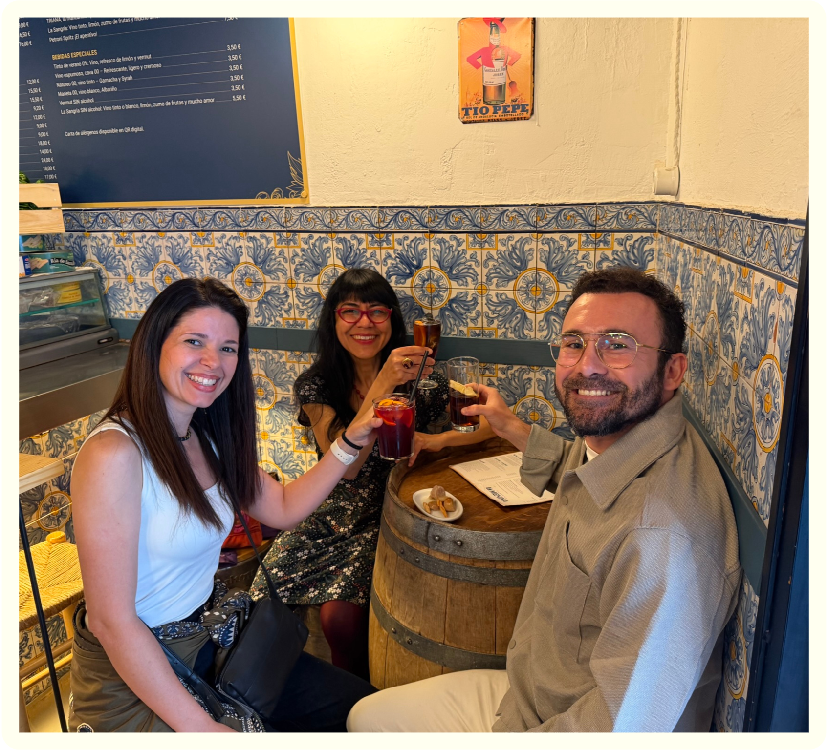 Three people sitting around a small round table in a restaurant, smiling and holding drinks. Two women and one man, with a colorful tile wall behind them.