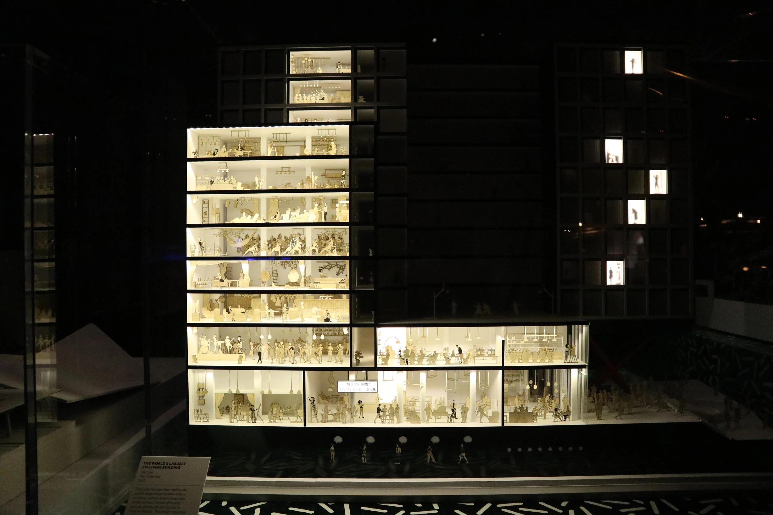 A detailed model of a multi-story building showcasing various interior scenes with miniature people engaging in different activities, illuminated from within, resembling a miniature city or complex.