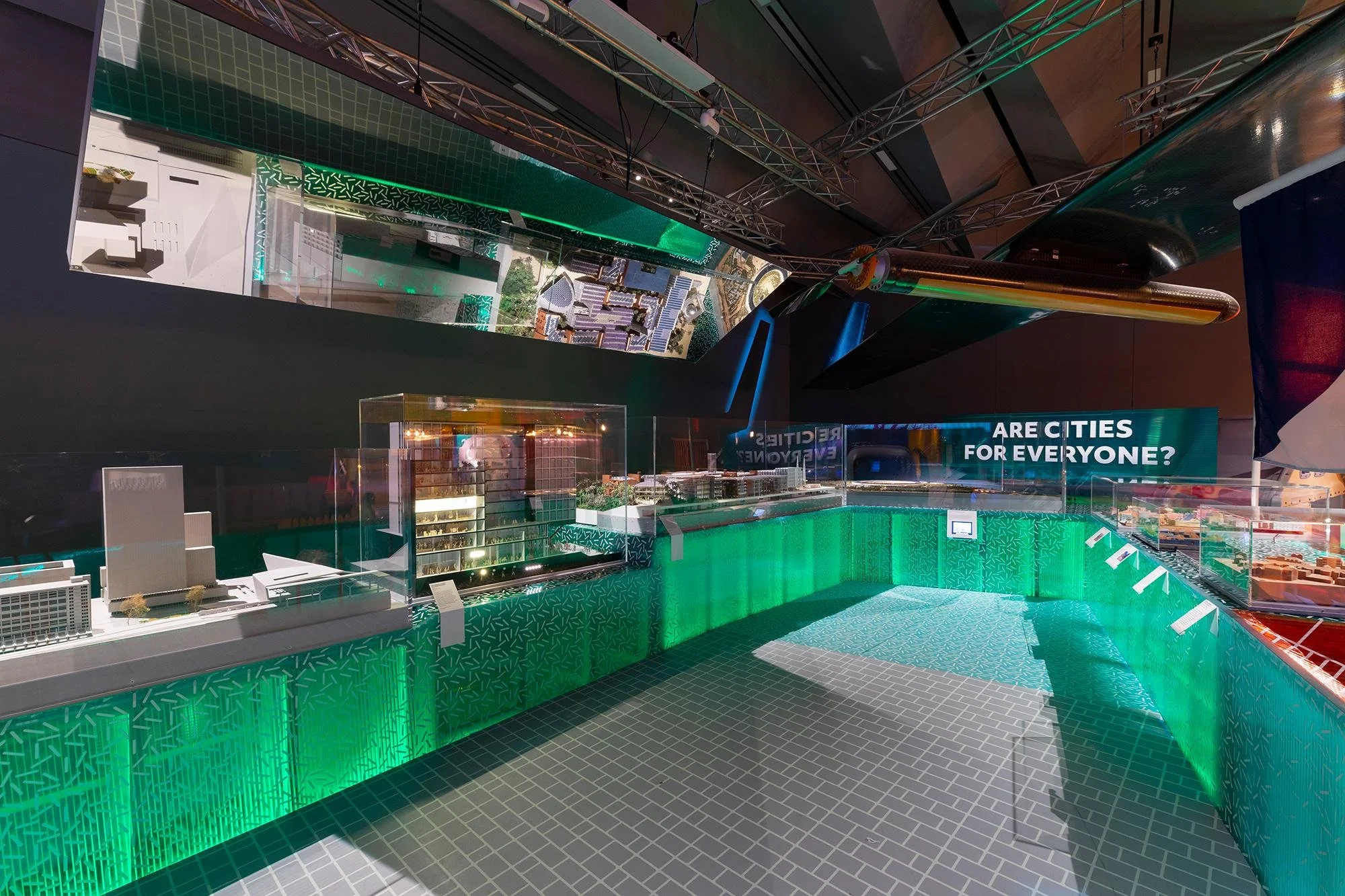 Interior view of a modern building with green LED lighting, model cityscape displays, and a question sign asking if cities are for everyone.