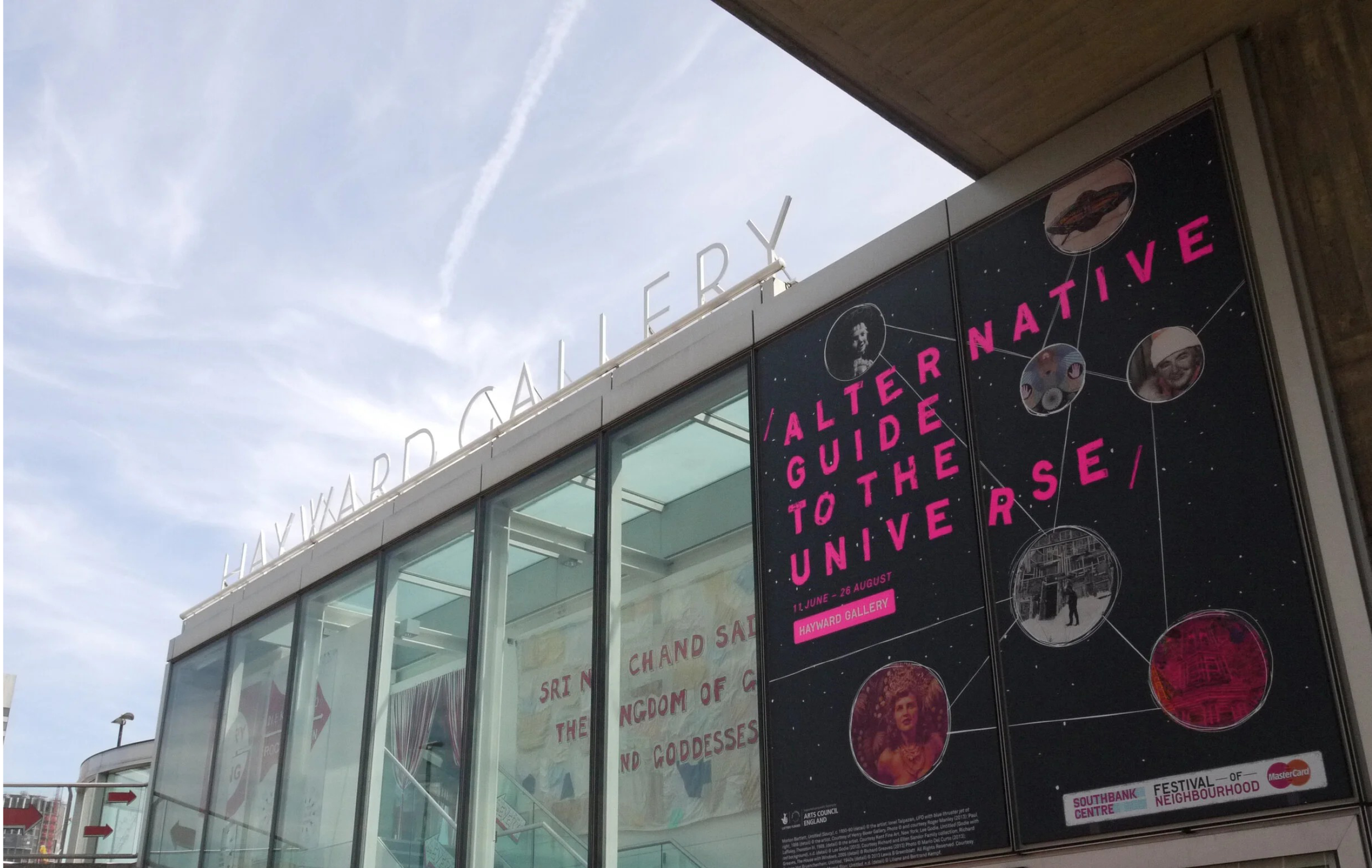 Poster advertising the 'Alter-Native Guide to the Universe' exhibition at Hayward Gallery, with images and text in pink on a black background, and a museum sign reading 'HAYWARD GALLERY' at the top.