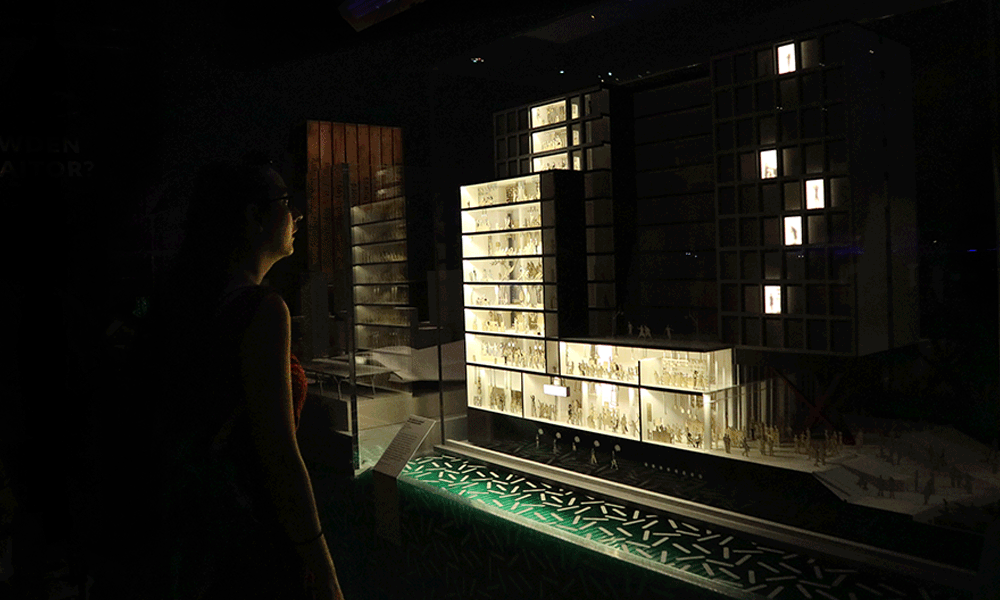 A woman looking at a detailed architectural model of a large multi-story building displayed in a dark room.