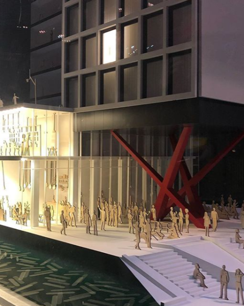 Architectural model of a modern building entrance with a large red sculpture and numerous miniature people figures.