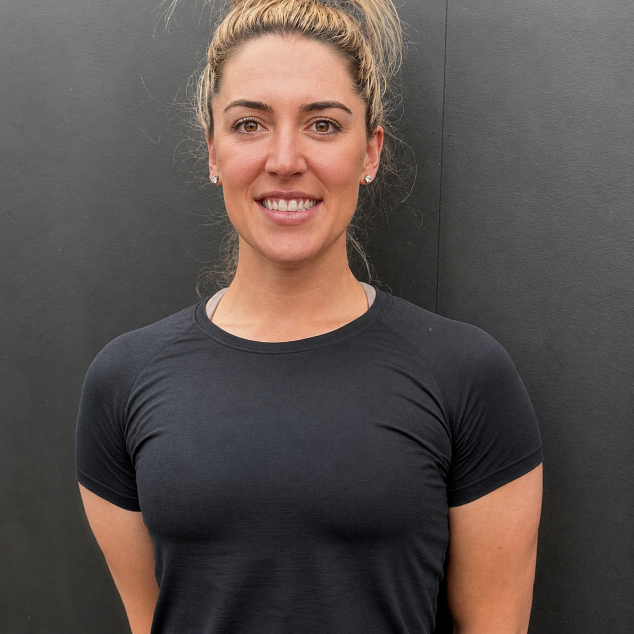 A woman with blonde hair tied up, wearing a black athletic shirt, standing against a dark wall, smiling at the camera.