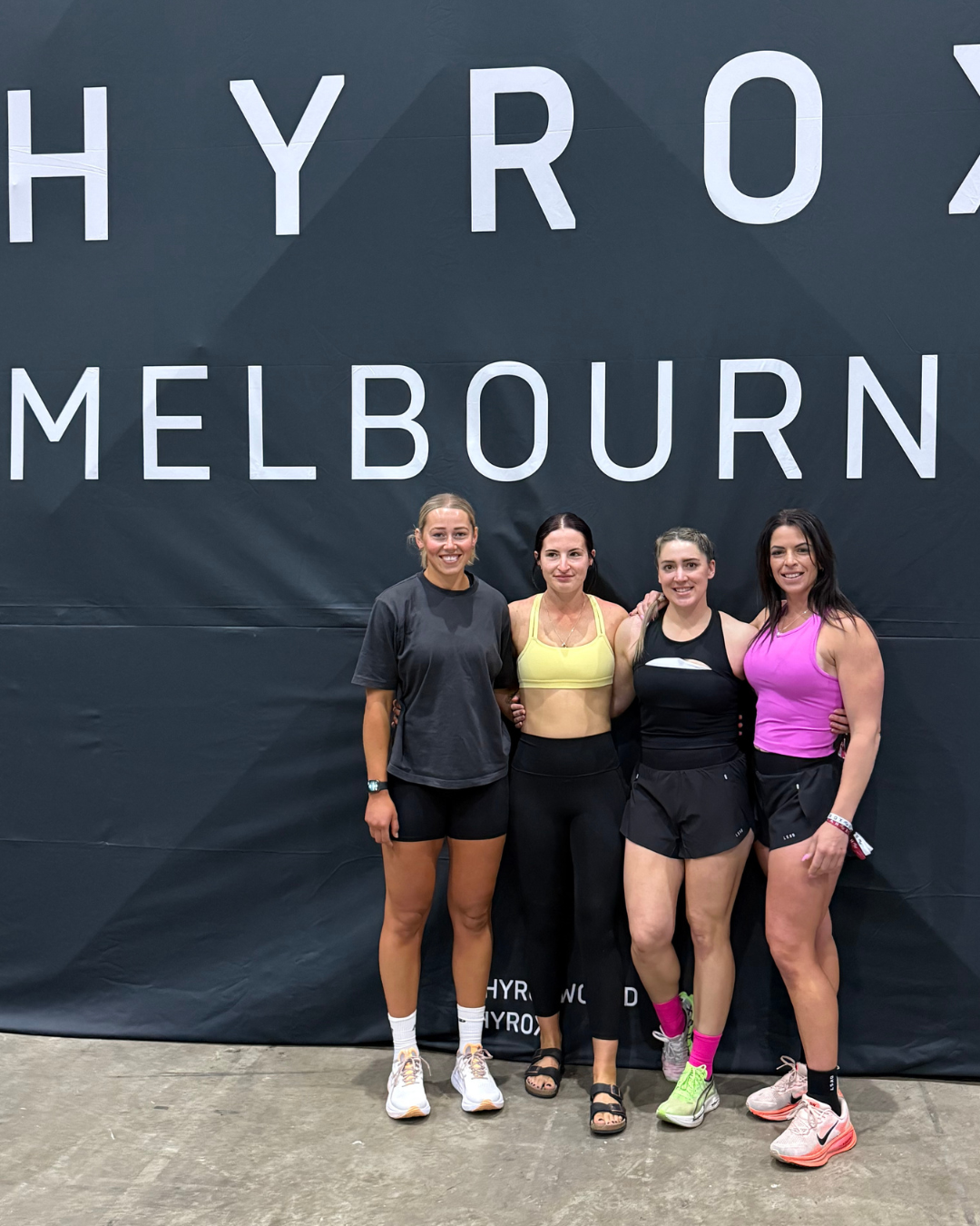 Four women standing in front of a large sign that reads 'HYROX MELBOURNE,' dressed in athletic wear, smiling after a fitness event.