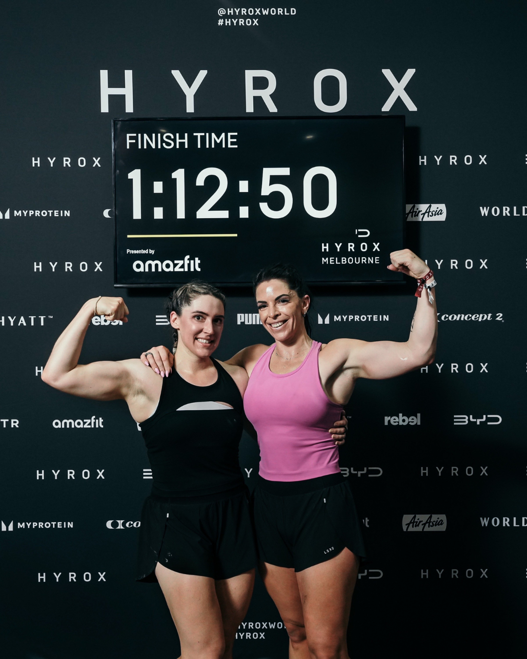 Two women at a fitness event, flexing muscles and smiling, in front of a finish time display showing 1:12:50 with the background branded with HYROX and and various sponsor logos.