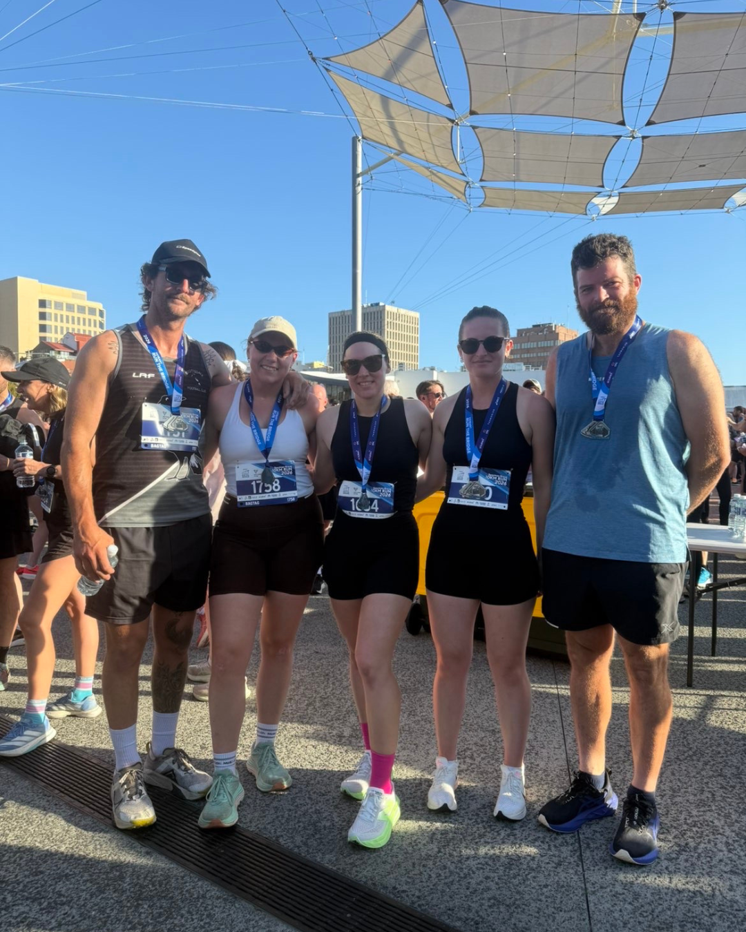 Five runners in athletic gear and medals standing outdoors after a race, with a cityscape and modern canopy structure above.