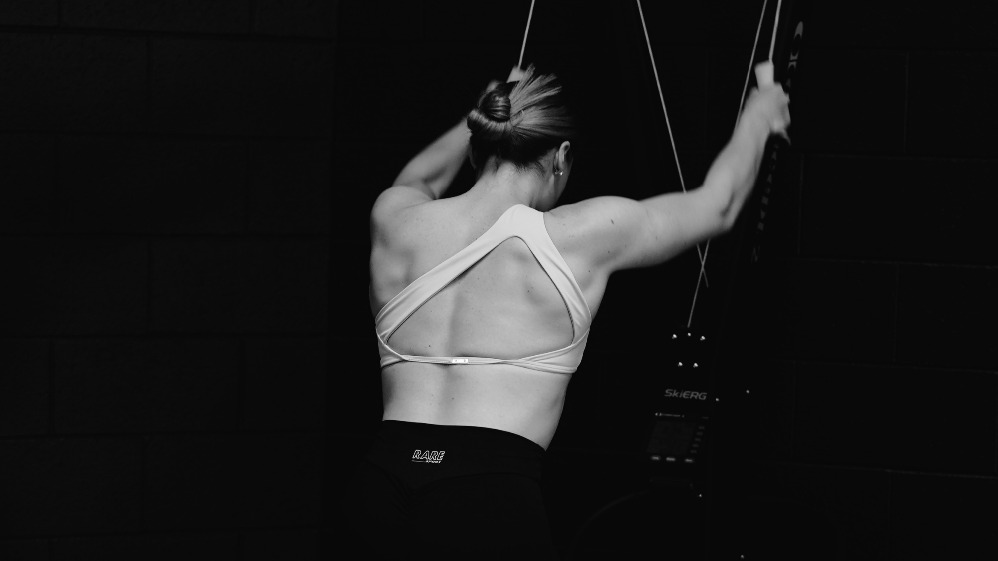 A woman with a bun hairstyle is elevating her arms while using a wall-mounted exercise machine in a gym. She is wearing a sports bra and shorts, with a dark background and a focus on her back muscles.