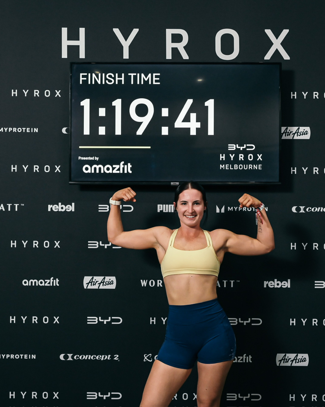 Female athlete smiling and flexing her biceps at the finish line of a race, displaying her muscular arms, with a large digital timer showing 1:19:41 behind her on a black backdrop with various sponsor logos.