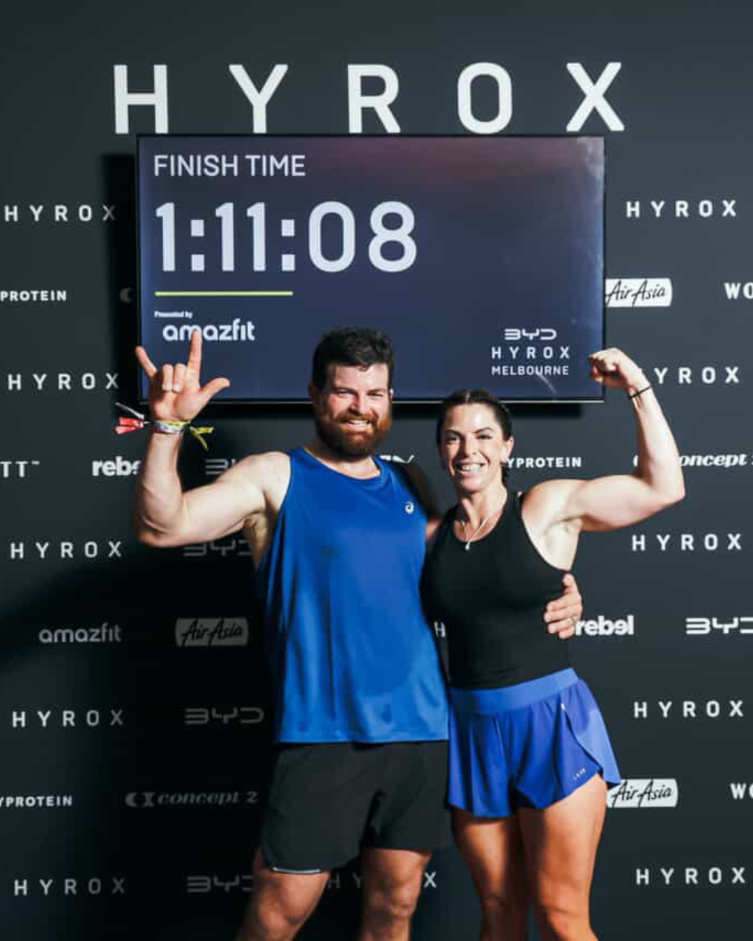Two fitness athletes smiling and posing for a photo after a race. The man is wearing a blue tank top and black shorts, flexing his right arm. The woman is wearing a black top and a blue skirt, flexing her left arm. They are standing in front of a black backdrop with words like 'HYROX', 'Amazfit', and 'AirAsia.' A digital clock above them shows their race finish time of 1:11:08.