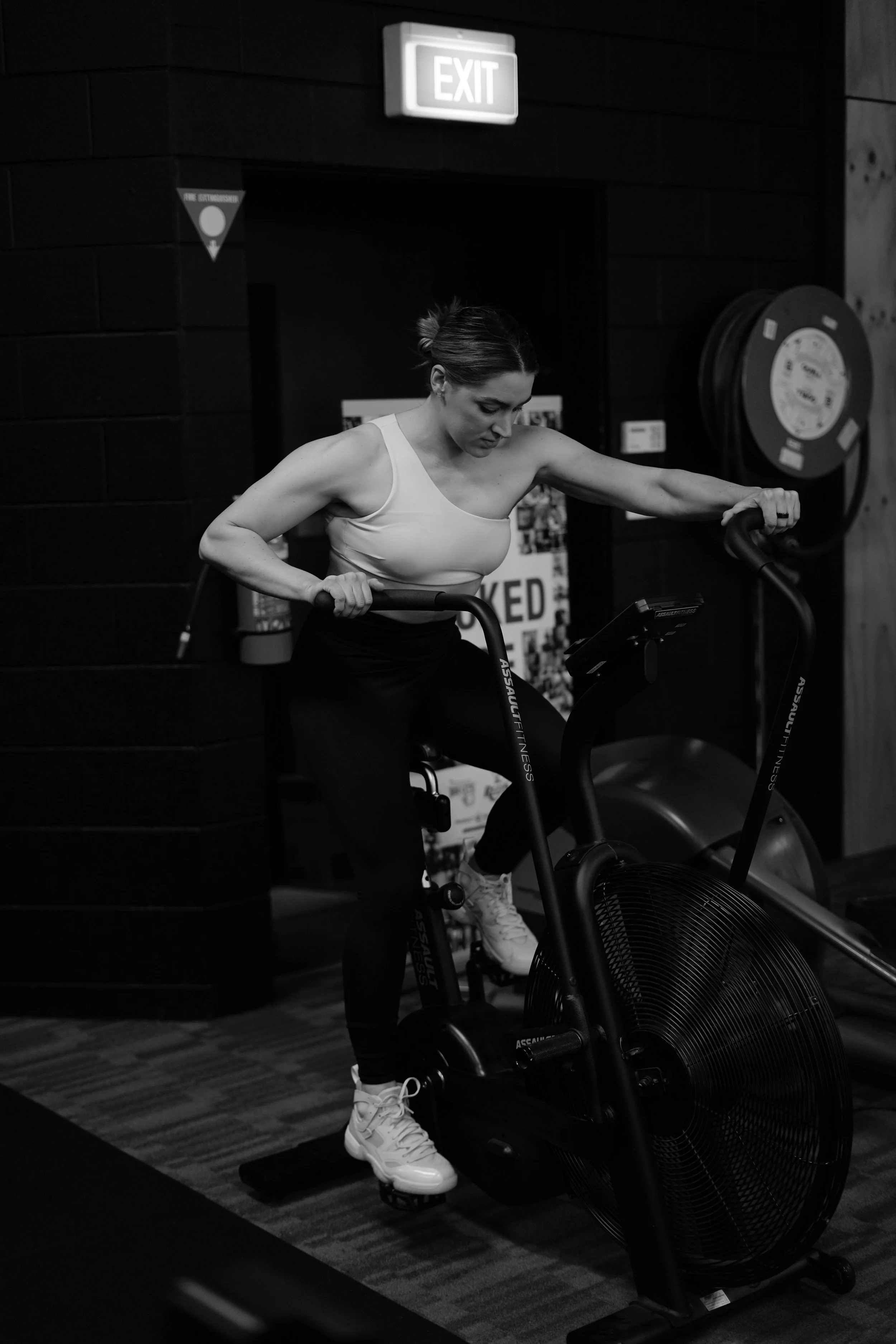 A woman in workout clothes exercising on a stationary bike in a gym, with an exit sign above her and gym equipment behind her.