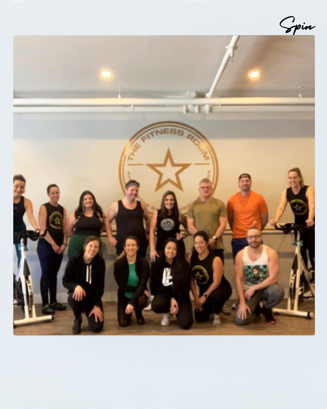 Group photo of fitness instructors and clients in a gym, with a logo that says 'The Fitness Room' and a star on the wall behind them, some people standing and some kneeling, with exercise bikes on either side.