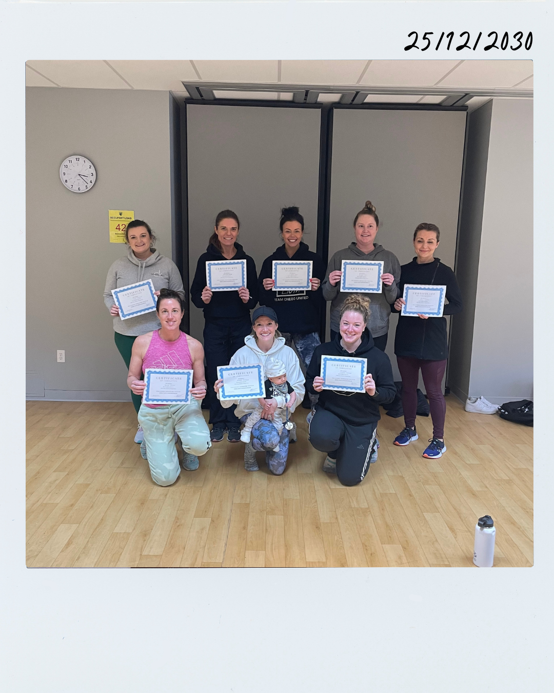 Group of nine women and one baby in athletic clothing holding certificates in a gym or fitness studio.