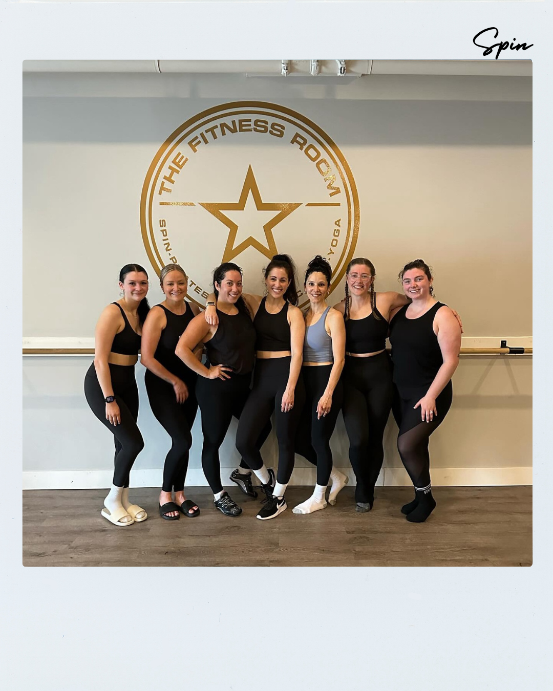 Seven women in workout clothes standing together in a fitness studio, smiling, with a large logo reading 'The Fitness Room' and a star on the wall behind them.