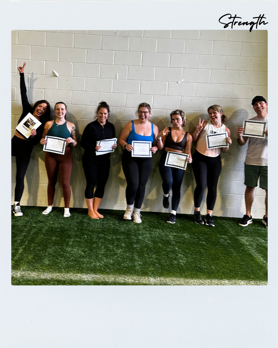 Group of seven smiling women and one man standing against a beige wall, holding certificates of achievement, some making playful gestures.