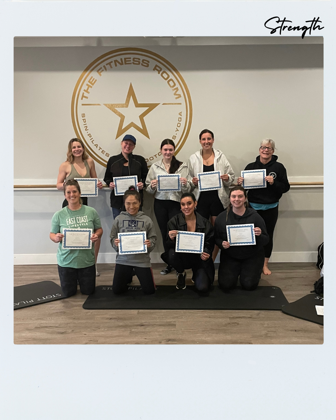 Group of women holding certificates in a fitness studio, standing in front of a wall with a large logo for 'The Fitness Room' and the words 'Strength' written in the corner.