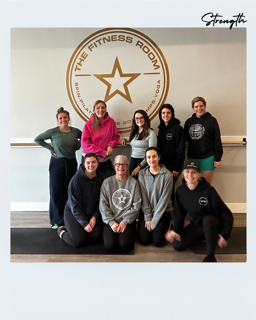 Group of nine women in workout attire posing in front of a wall with a The Fitness Room logo, some kneeling and some standing, inside a gym.