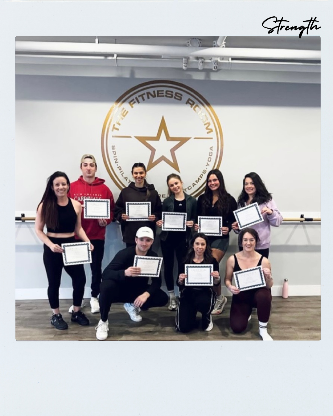 Group of ten young adults holding certificates in a fitness studio with a logo that reads 'The Fitness Room' and a star graphic on the wall, some people kneeling in the front row and others standing behind.