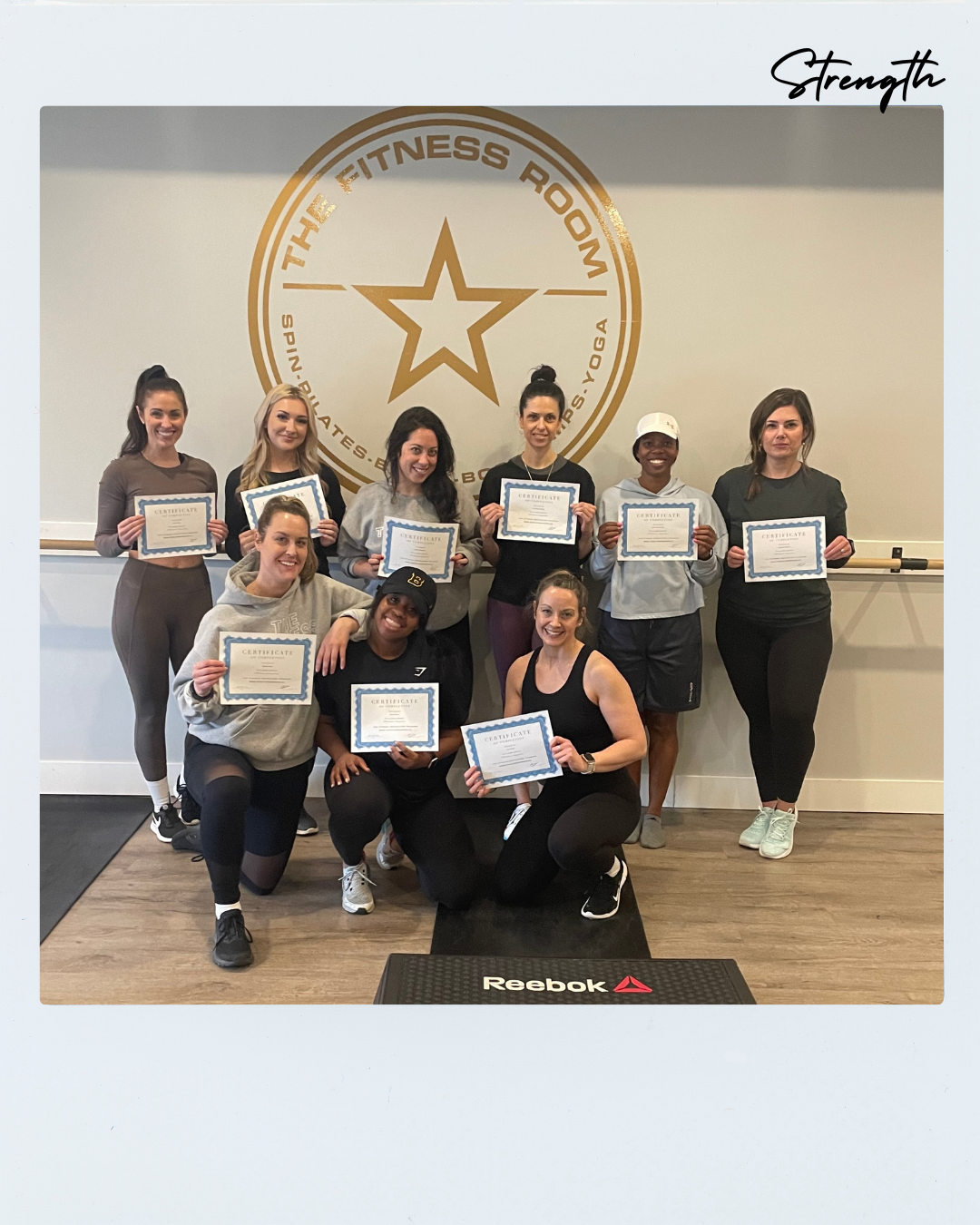 Group of women in a fitness studio holding certificates, standing and kneeling in front of a wall with a gold logo of the fitness room that features a star and circular text, with the word 'Strength' written in the top right corner.