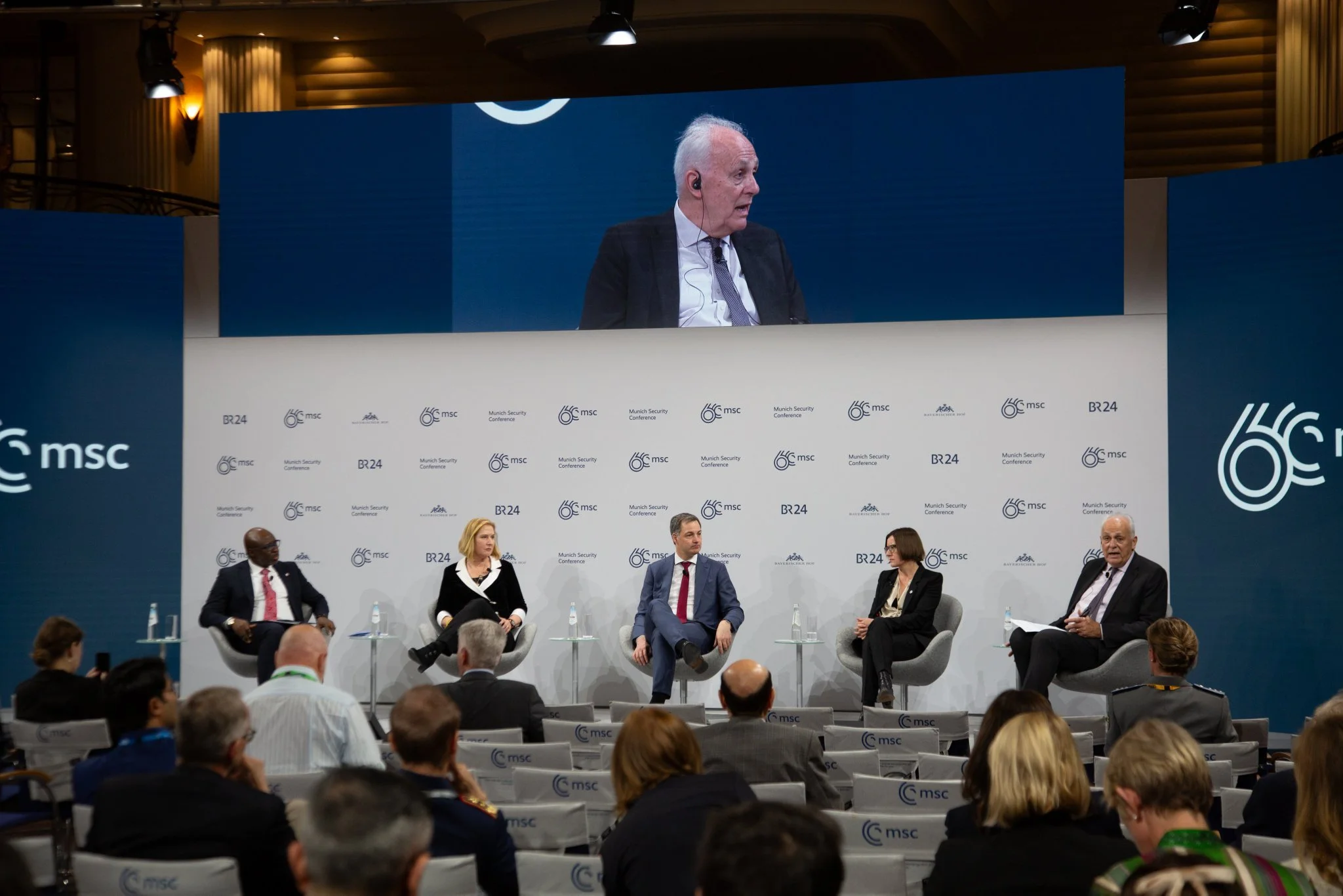 A panel discussion at the Munich Security Conference with five individuals seated on stage and a large screen displaying an elderly man speaking. The backdrop features the conference branding with multiple logos and the number 60.